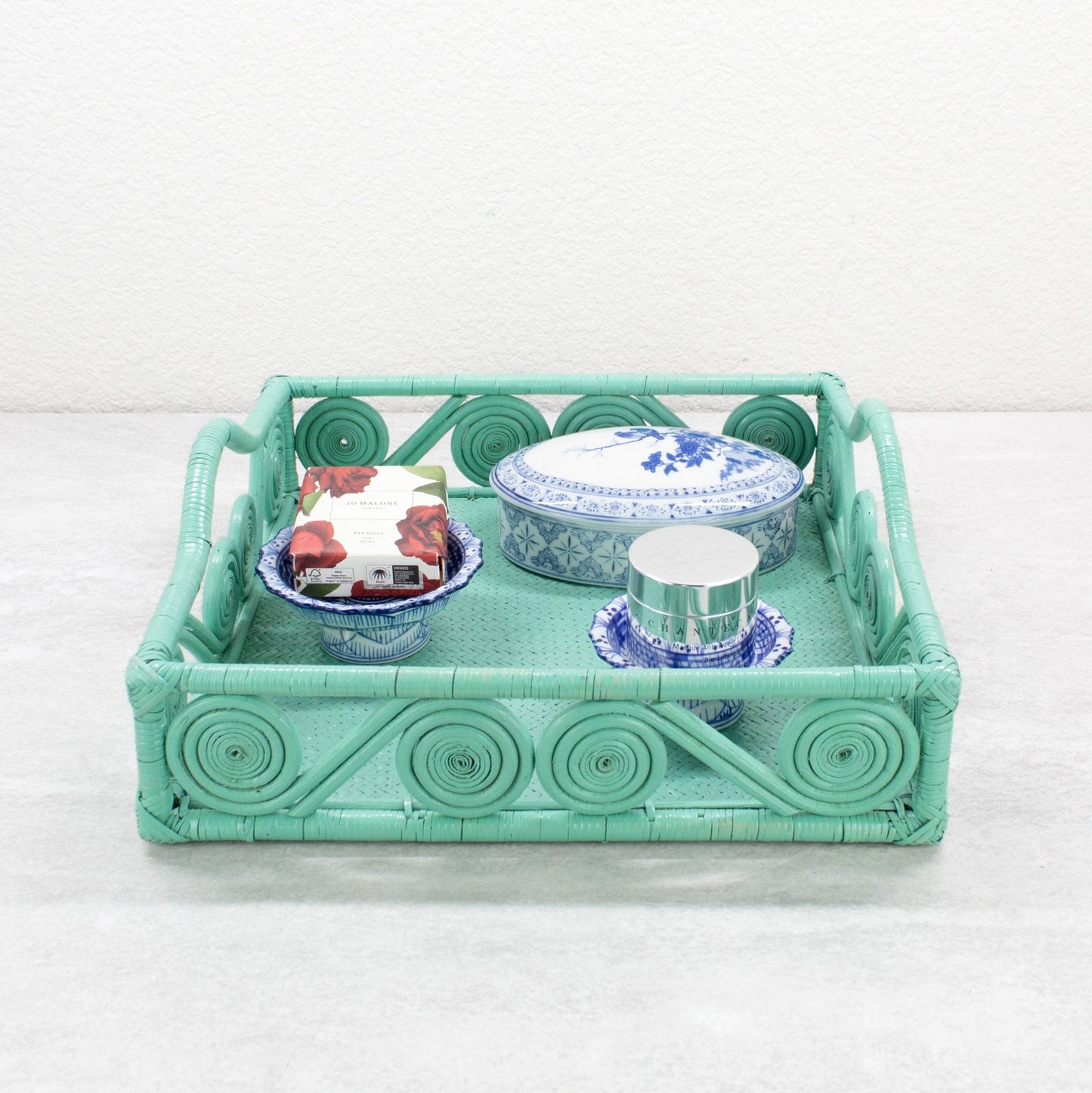 Turquoise rattan tray styled with blue porcelain dishes, a floral soap bar, and a silver cosmetic jar.