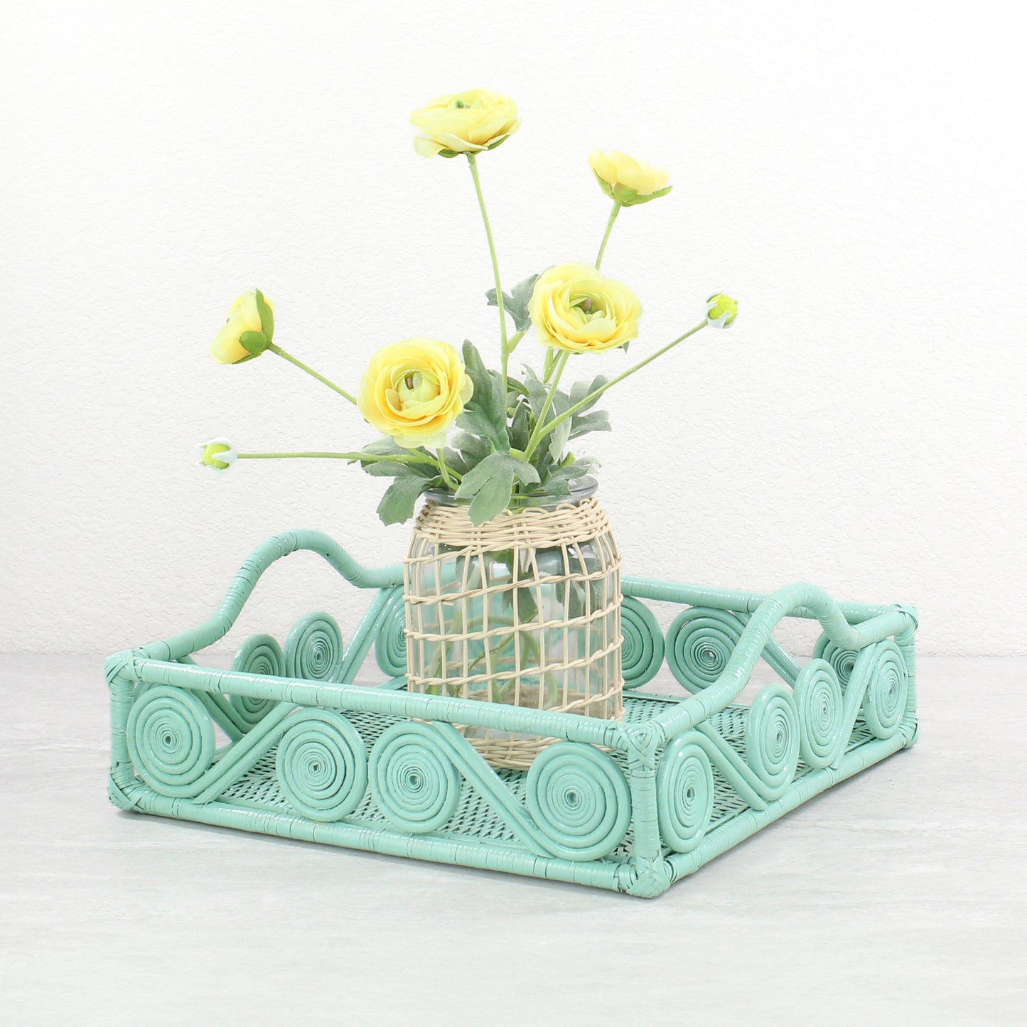 Sage leaf green rattan square tray styled with a woven vase of yellow ranunculus flowers on a neutral tabletop.