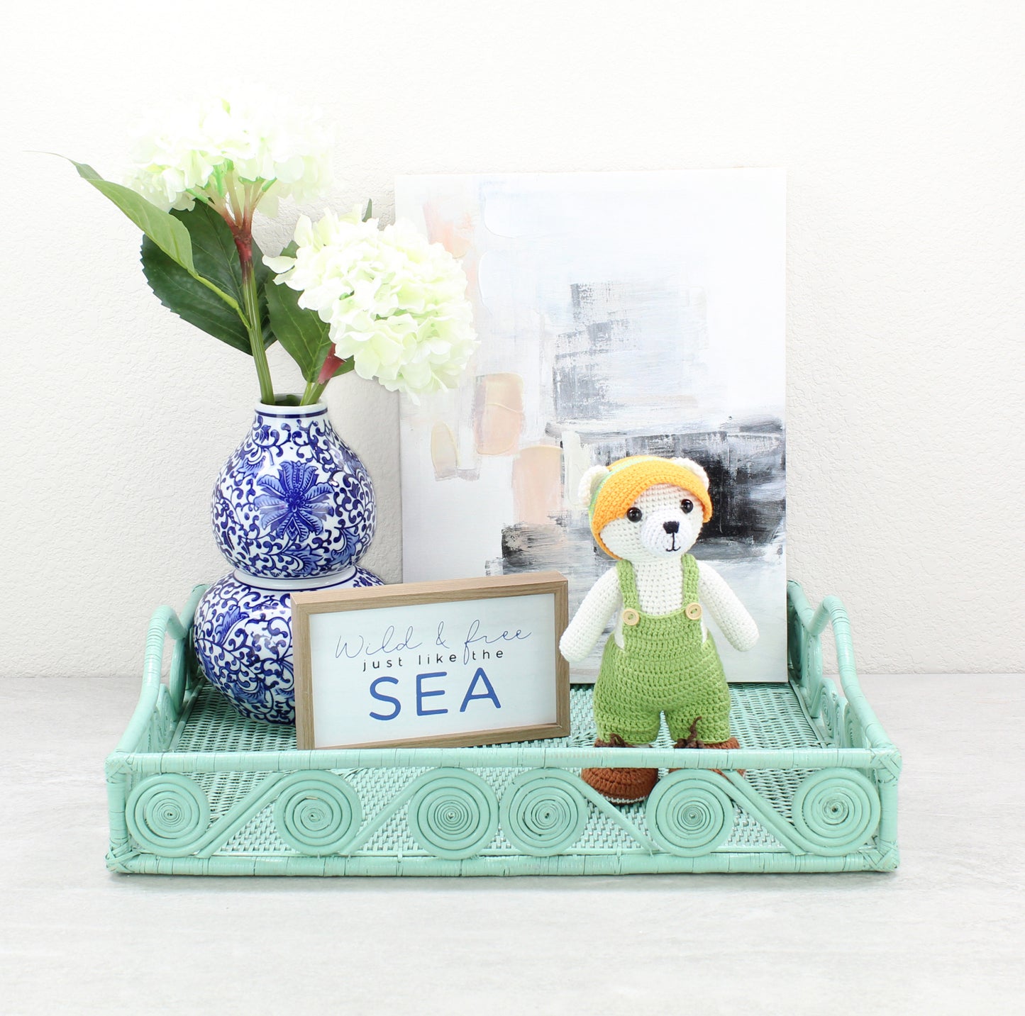 Sage leaf green rattan tray styled with hydrangeas in a blue vase, framed art, and handmade crochet bear décor.