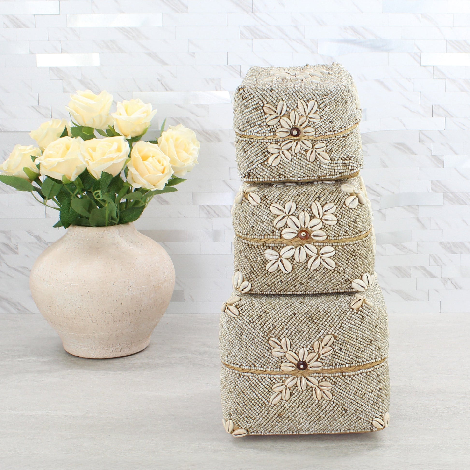 Stacked set of three handmade beige beaded cowrie shell boxes beside a ceramic vase, styled in a bright coastal-inspired setting.