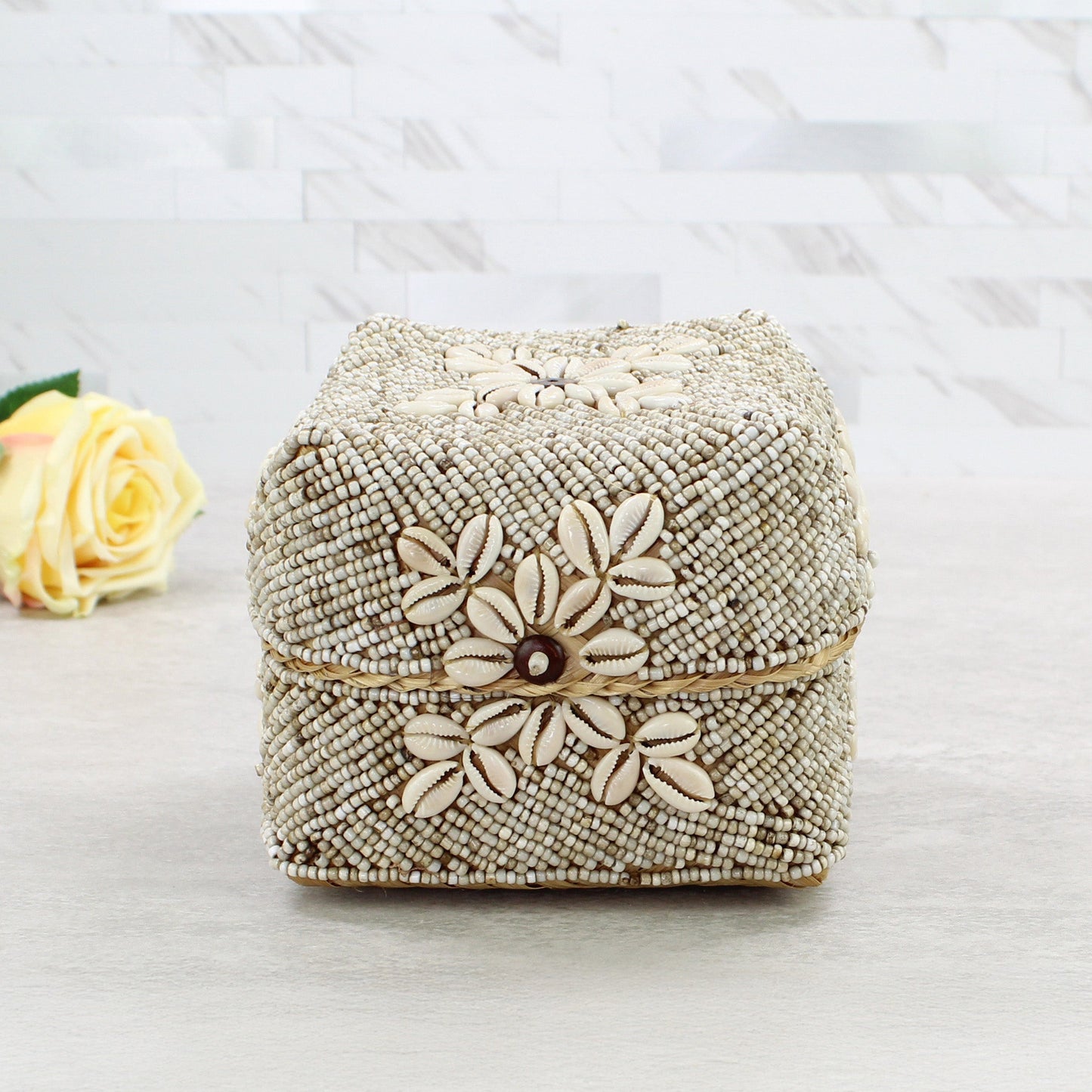 Small beige beaded storage box decorated with natural cowrie shells and central flower motif in coastal boho décor style.