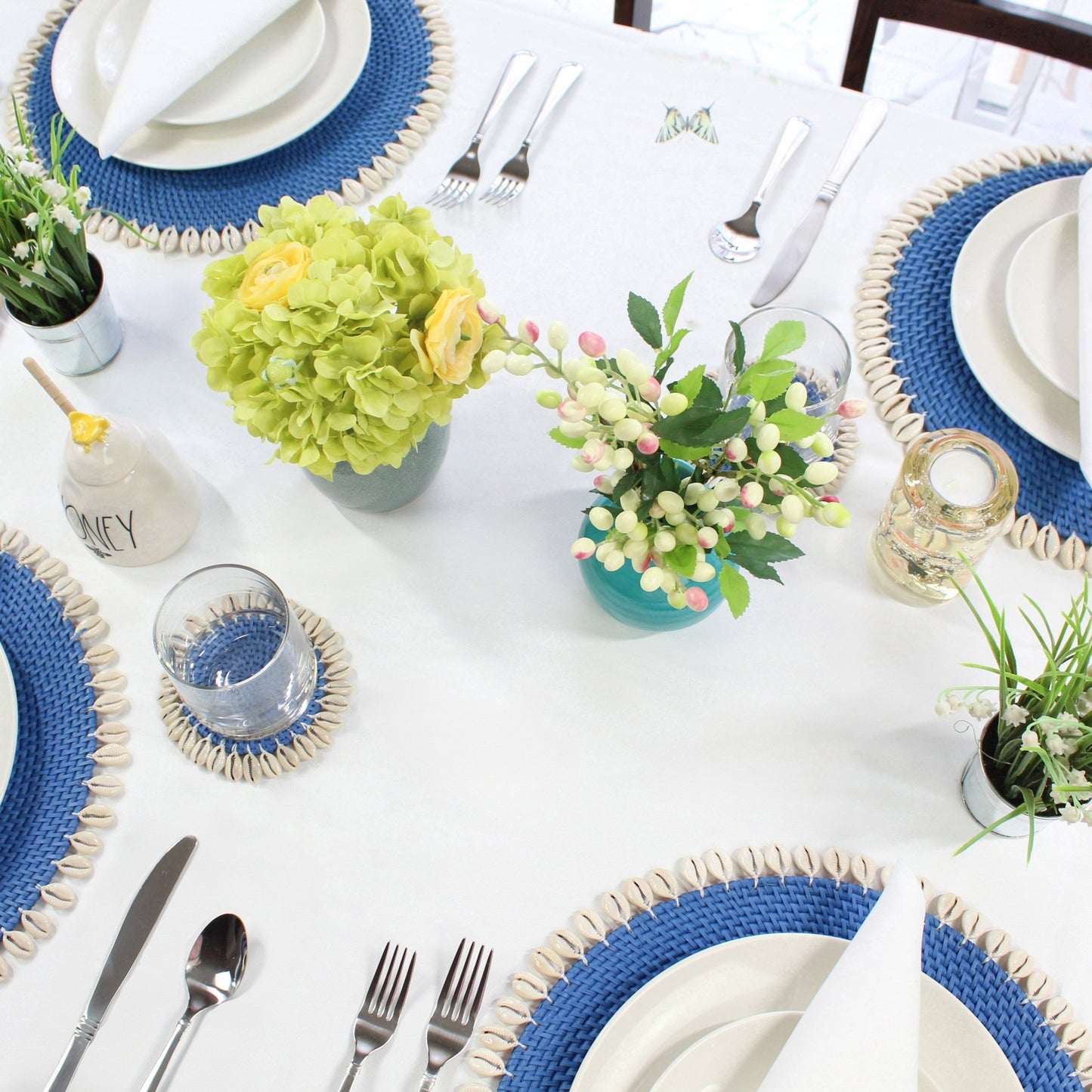 Coastal dining table styled with blue seashell-trimmed rattan placemats, white plates, floral centerpiece, and silver cutlery