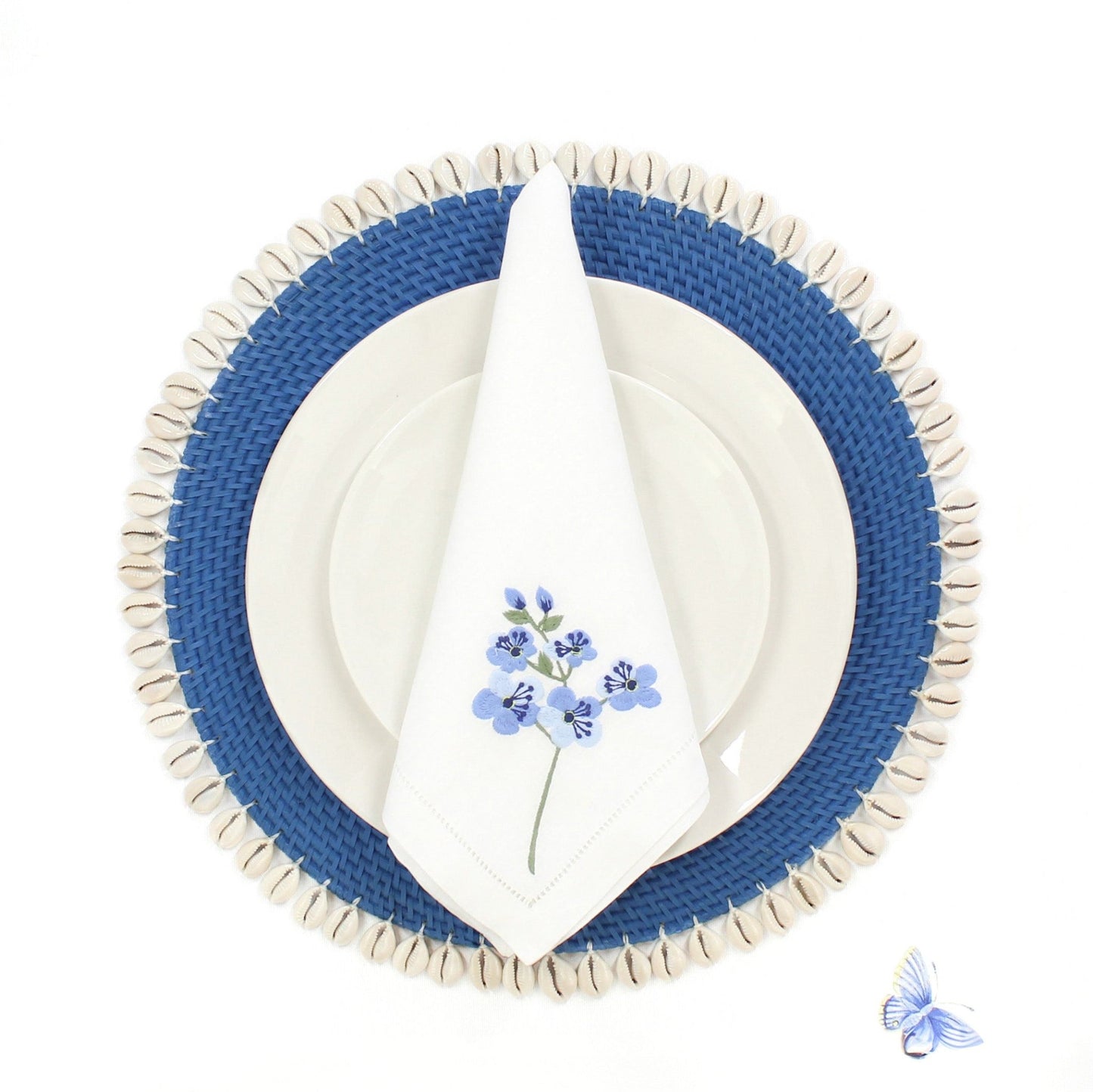 Blue rattan placemat with seashell trim, white plate, and floral napkin styled on a white dining table for coastal decor.