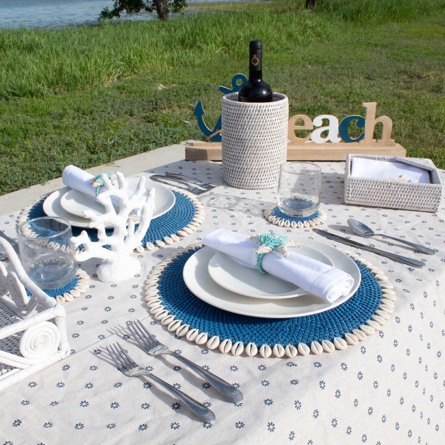 Outdoor coastal table set with blue rattan placemats, whitewash wine holder, flowers, and cutlery by the lake