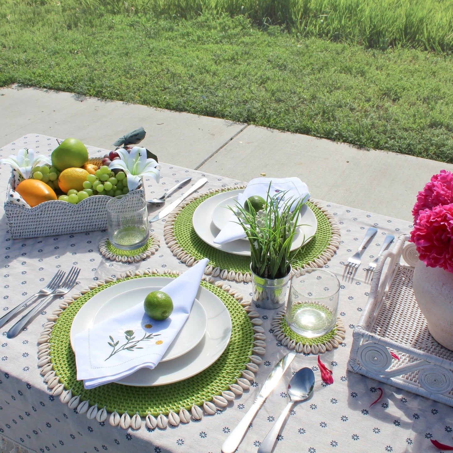 Lime green seashell-trimmed rattan placemats and coasters styled on an outdoor dining table with fruits, napkins, and flowers.