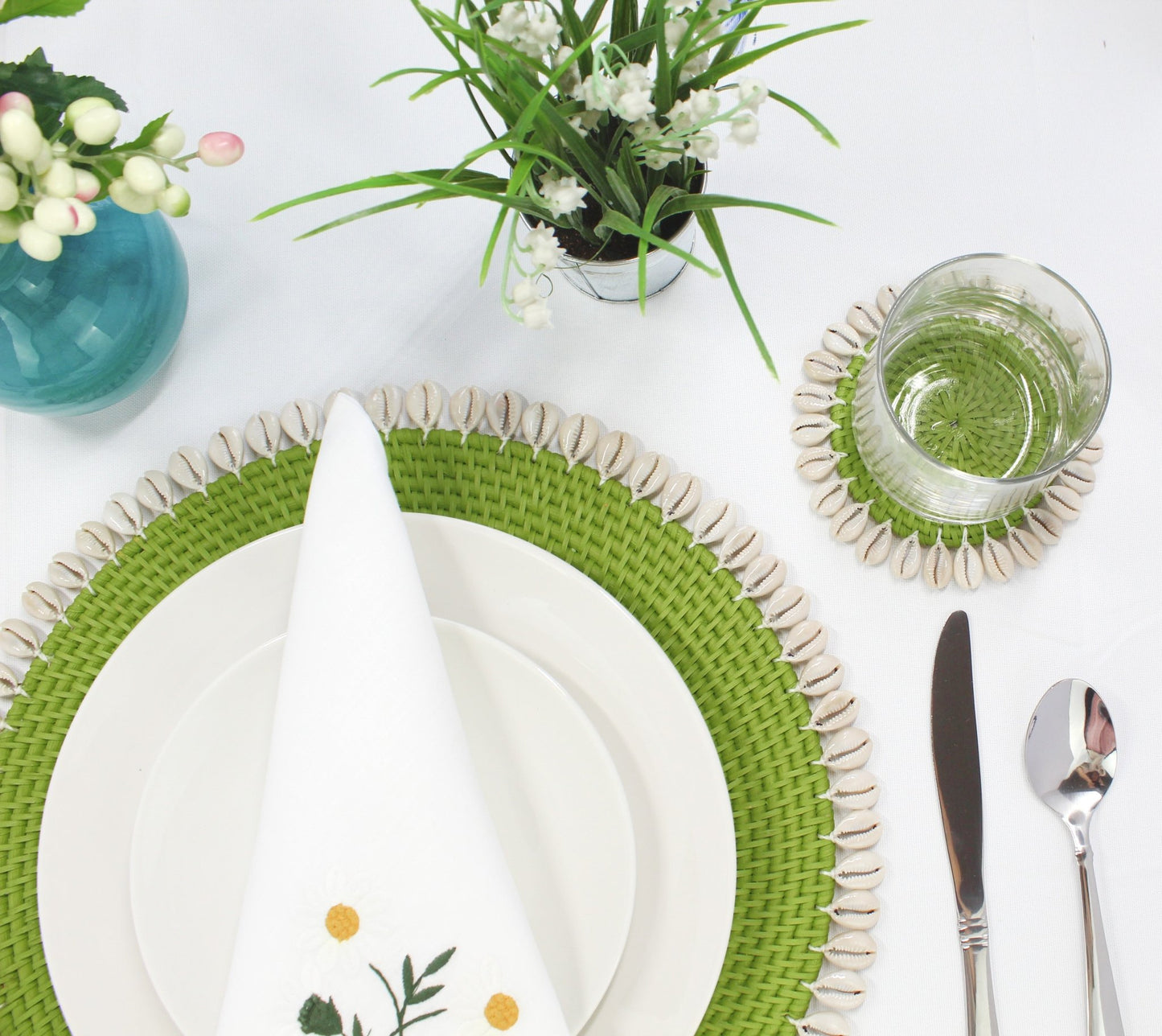 Green seashell-trimmed rattan placemat and matching coaster styled on a white dining table with fresh flowers and glassware