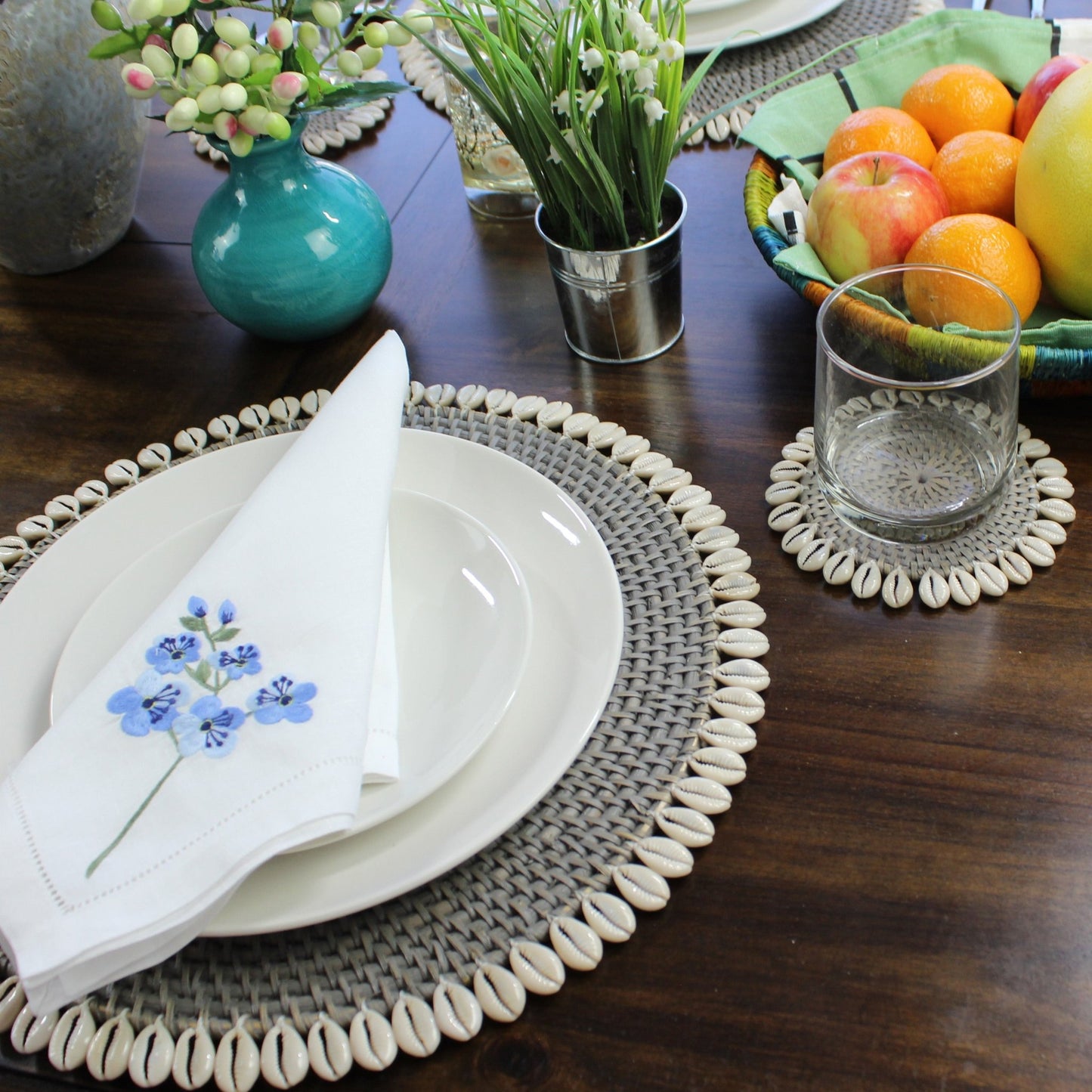 grey seashell-trimmed rattan placemat used as charger under dinnerware, protecting tabletop and enhancing coastal presentation
