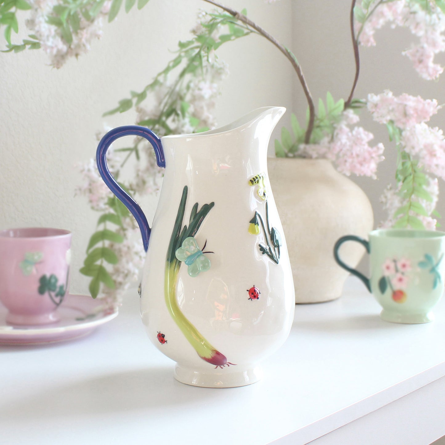 White stoneware pitcher with hand-painted garden-inspired motifs including flowers, butterflies, and ladybugs, artisan glazed ceramic jug