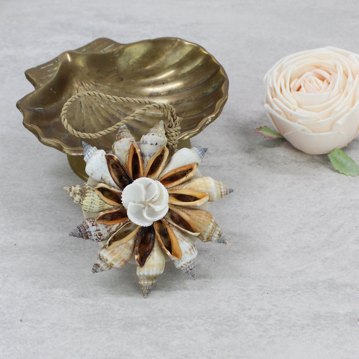 Natural Bali handmade shell Christmas bauble with spiral shells and central rose shell design, displayed beside brass bowl and soft pink flower on a stone table.
