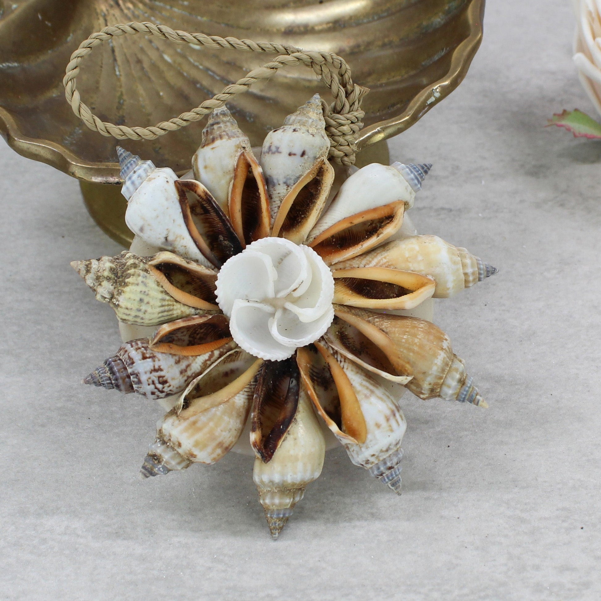 Natural Bali handmade shell Christmas bauble with spiral shells and central rose shell design, displayed beside brass bowl and soft pink flower.