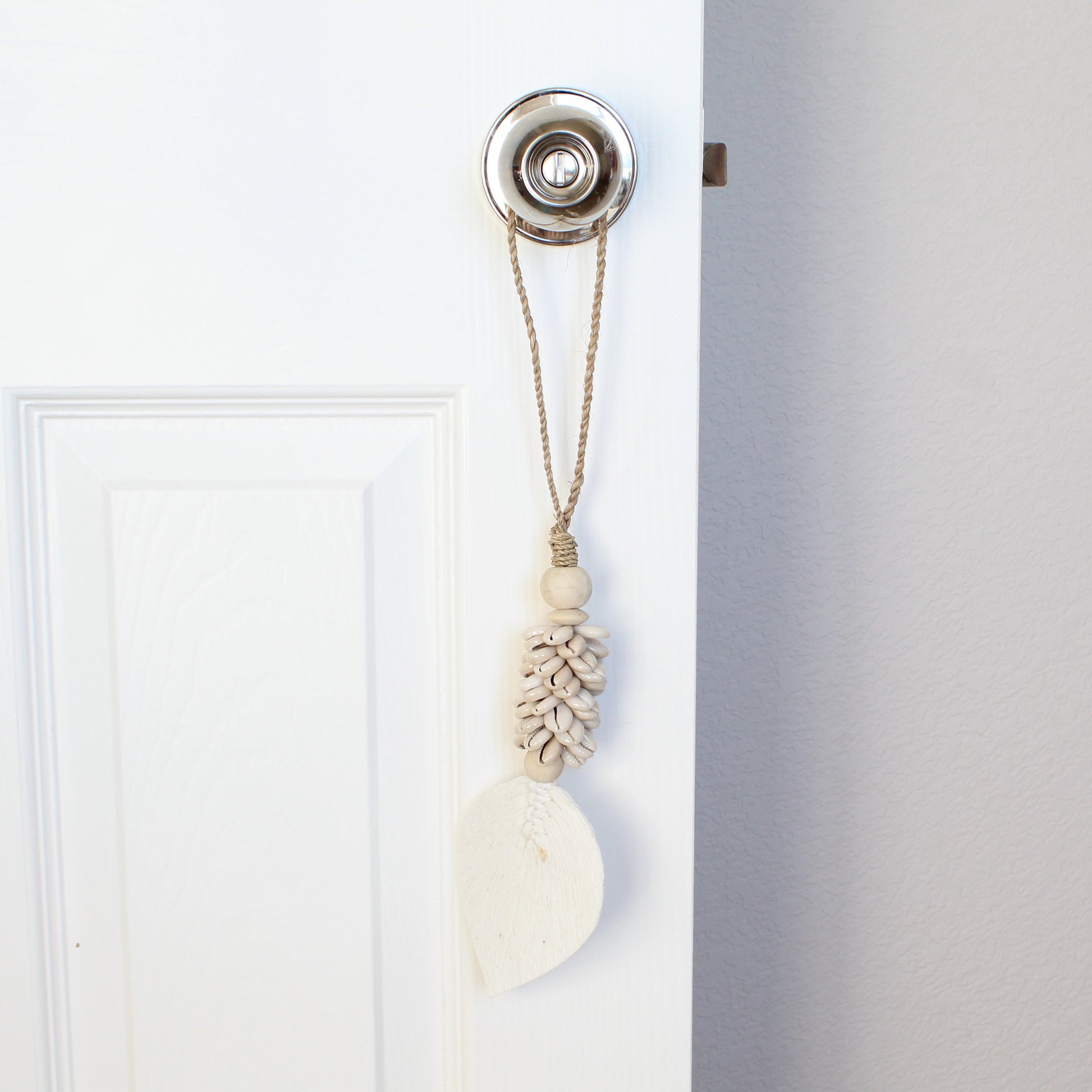 Beige beaded seashell tassel with braided jute loop used as boho curtain tieback on white door handle