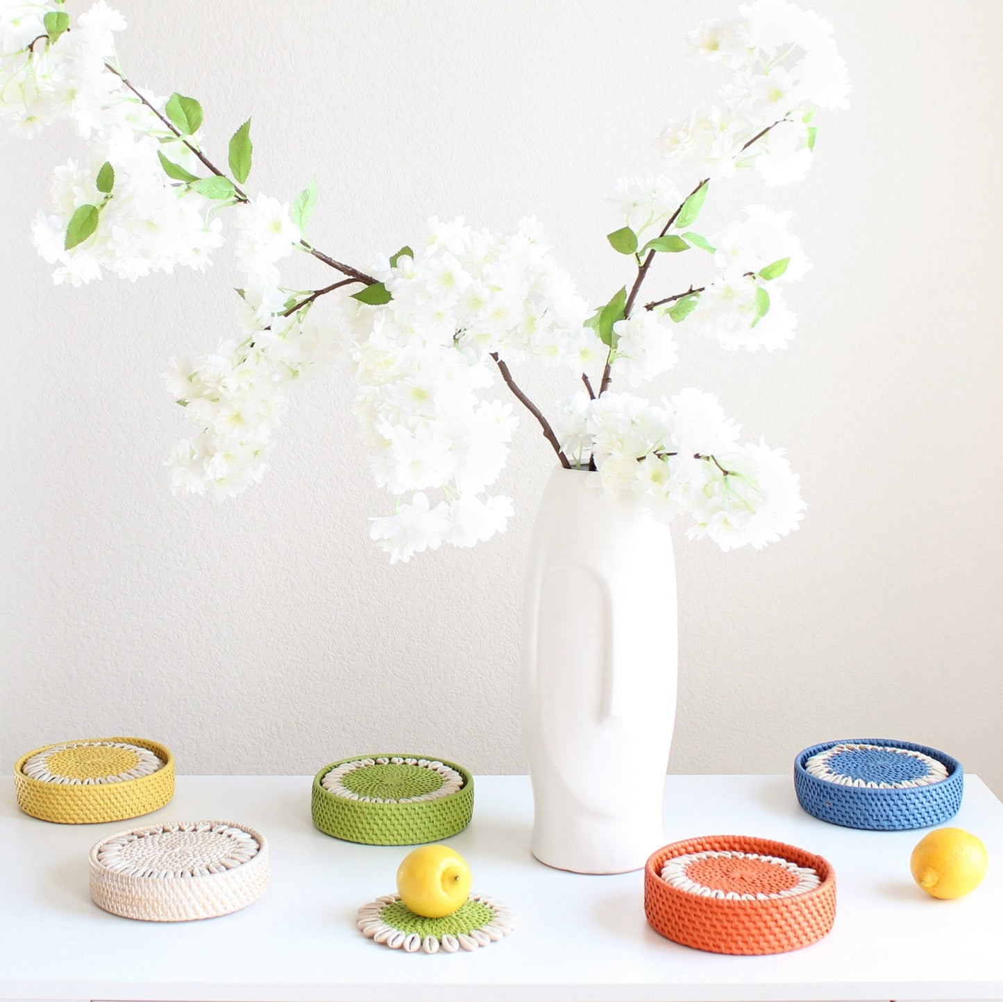 Colorful rattan coasters with seashell trim styled near vase with flowers and citrus fruits for vibrant coastal table décor.