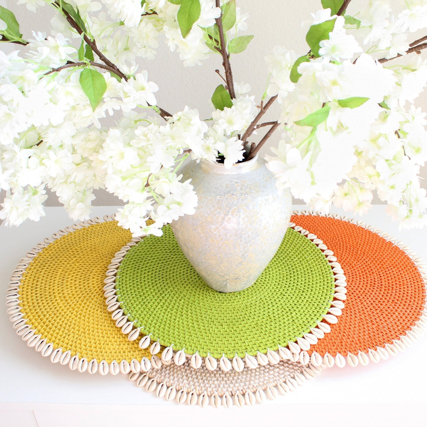 Seashell-trimmed rattan placemats shown in whitewash, gray, yellow, lime green, orange, and blue