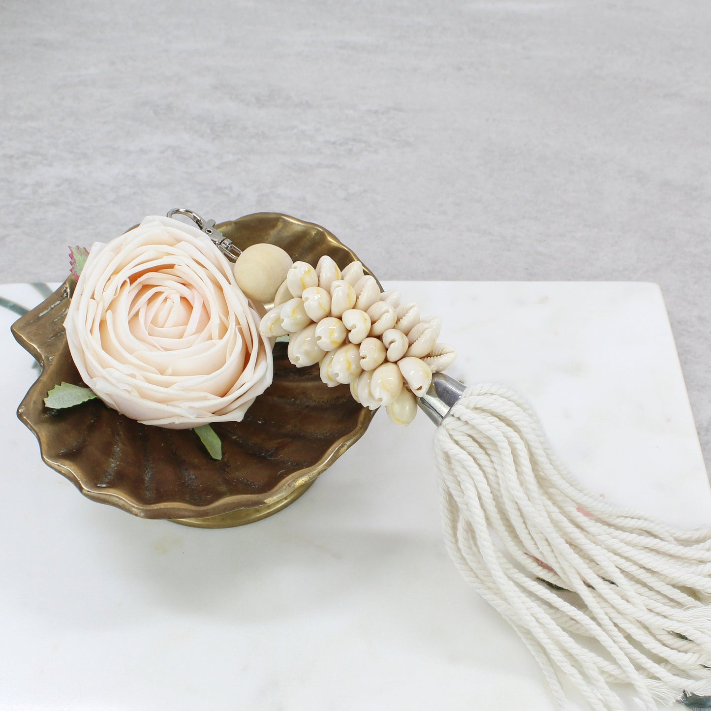 Natural sand seashell key ring with white cotton tassel resting in shell-shaped brass dish beside blush rose accent.