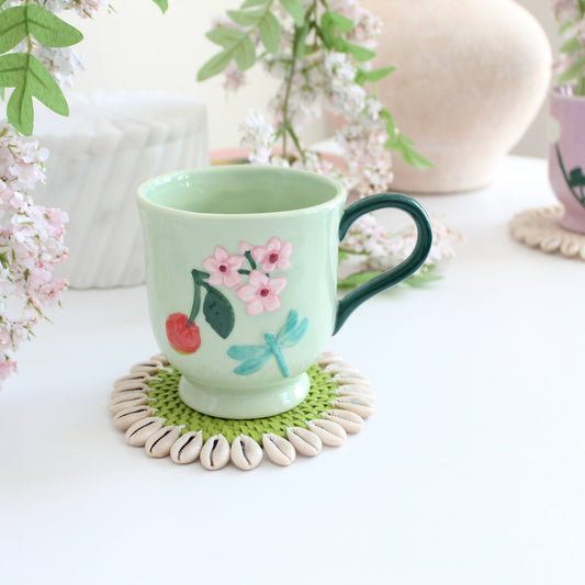 Lime green seashell-trimmed rattan coaster under ceramic mug on wooden table, protecting surface from heat and moisture