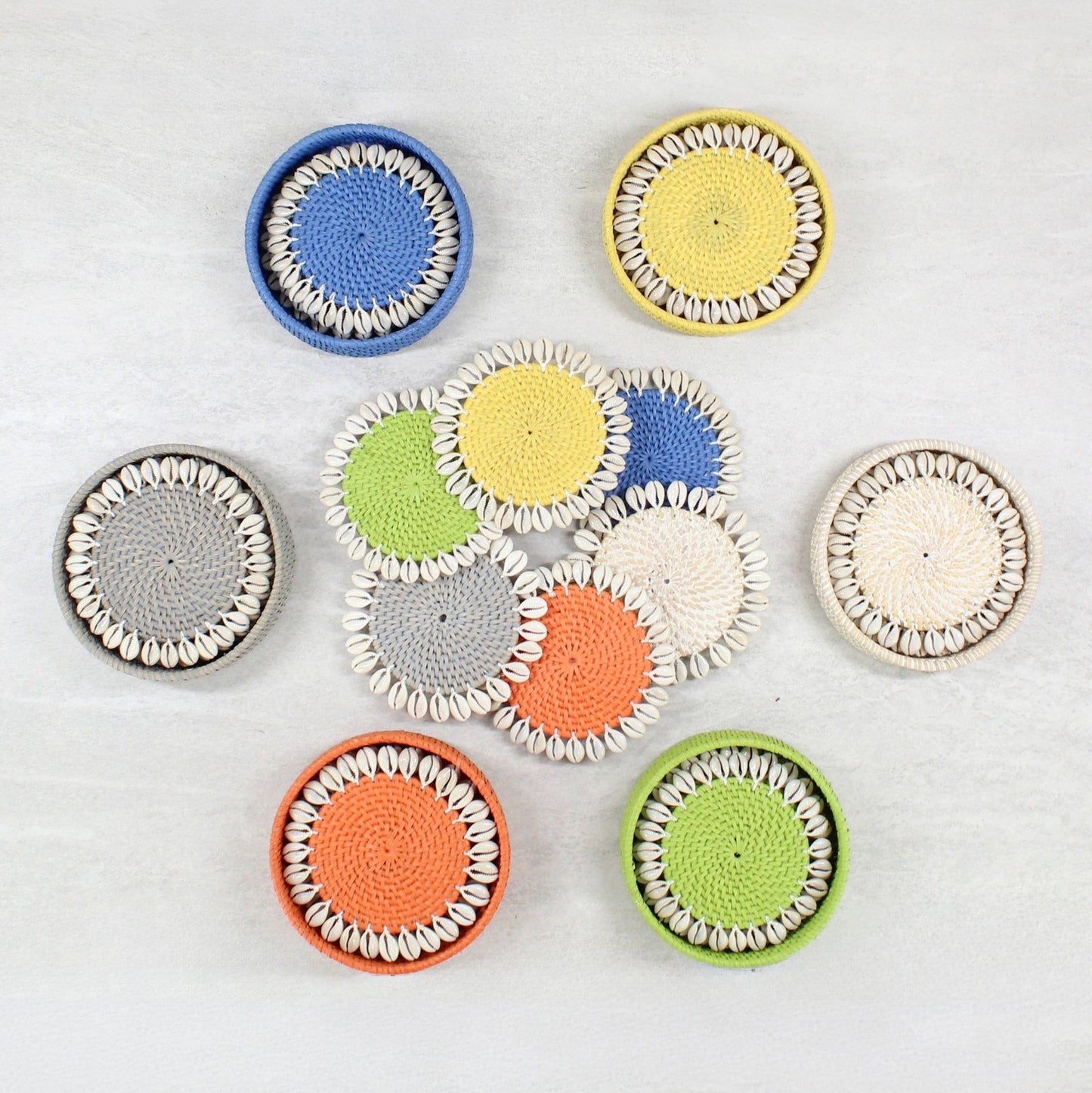 Seashell-trimmed rattan coasters in whitewash, gray, yellow, lime green, orange, and blue, with holders
on a stone table