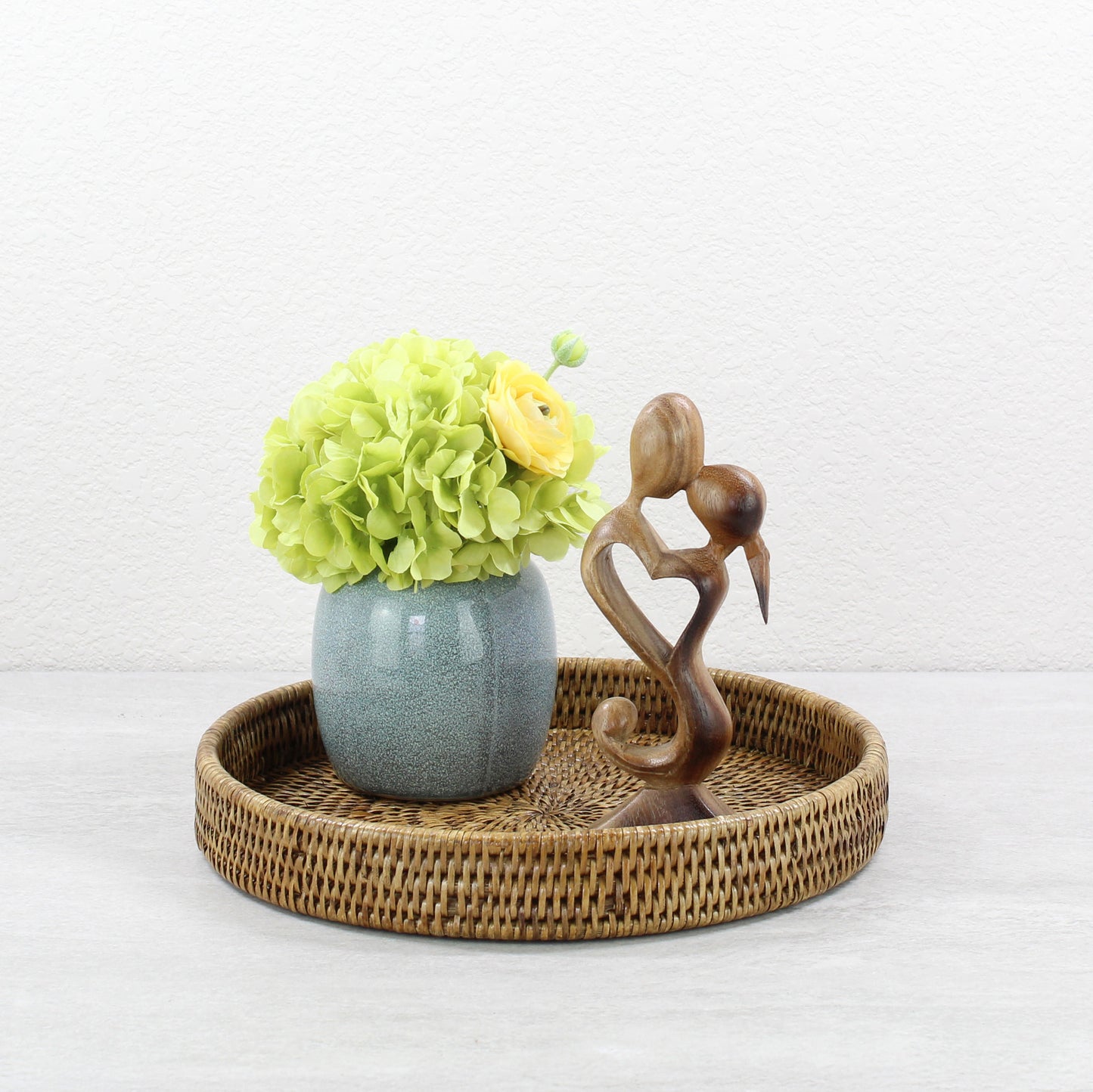 Decorative round honey brown rattan tray styled with a ceramic vase of flowers and a wooden couple sculpture, eco-friendly home décor inspiration.