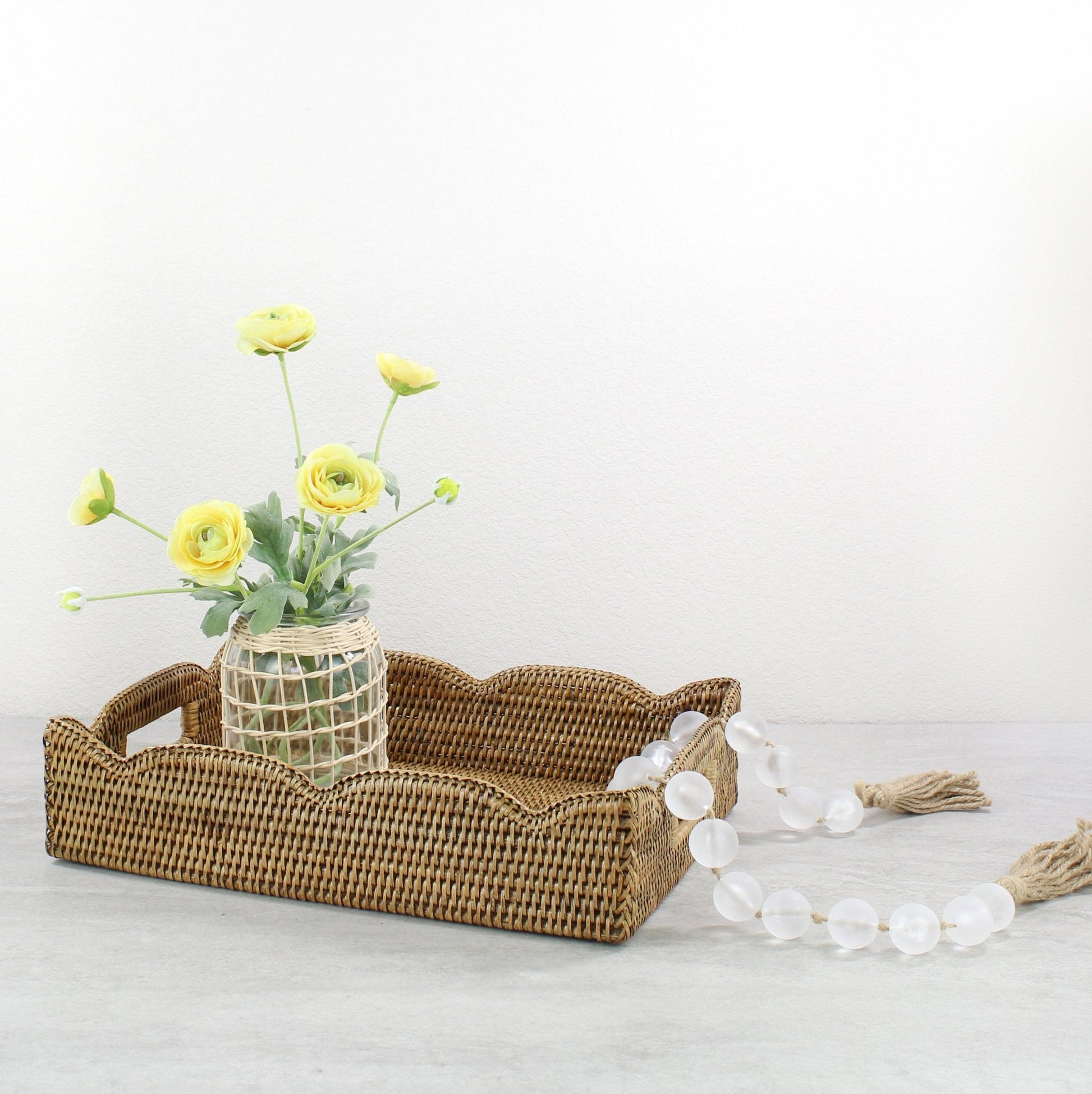 Scalloped rattan tray in honey brown styled with woven vase of yellow flowers and glass bead garland, handmade eco-friendly decorative accent.