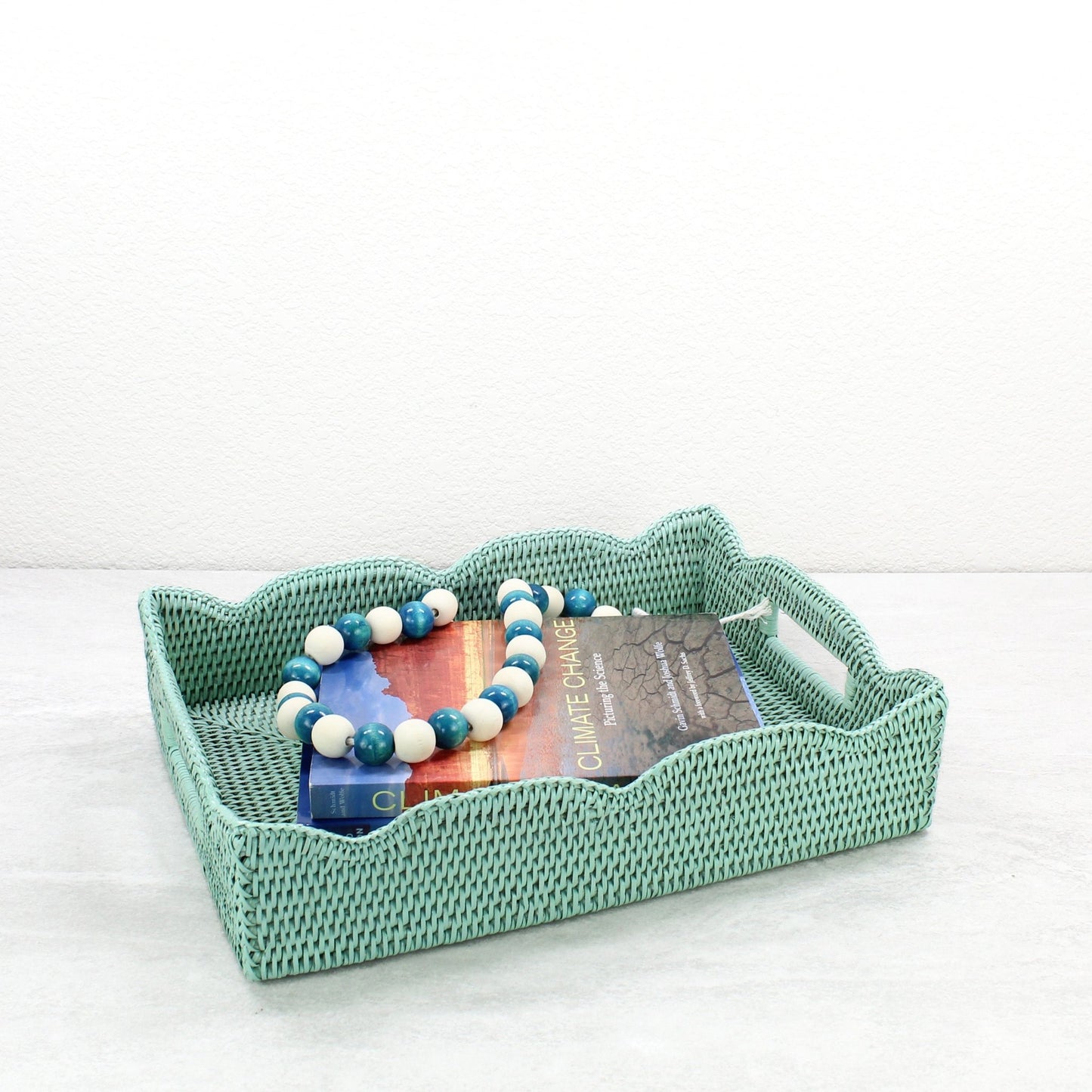 Sage Leaf Green scalloped rattan tray styled with a climate change book and blue-white bead garland on a light tabletop.