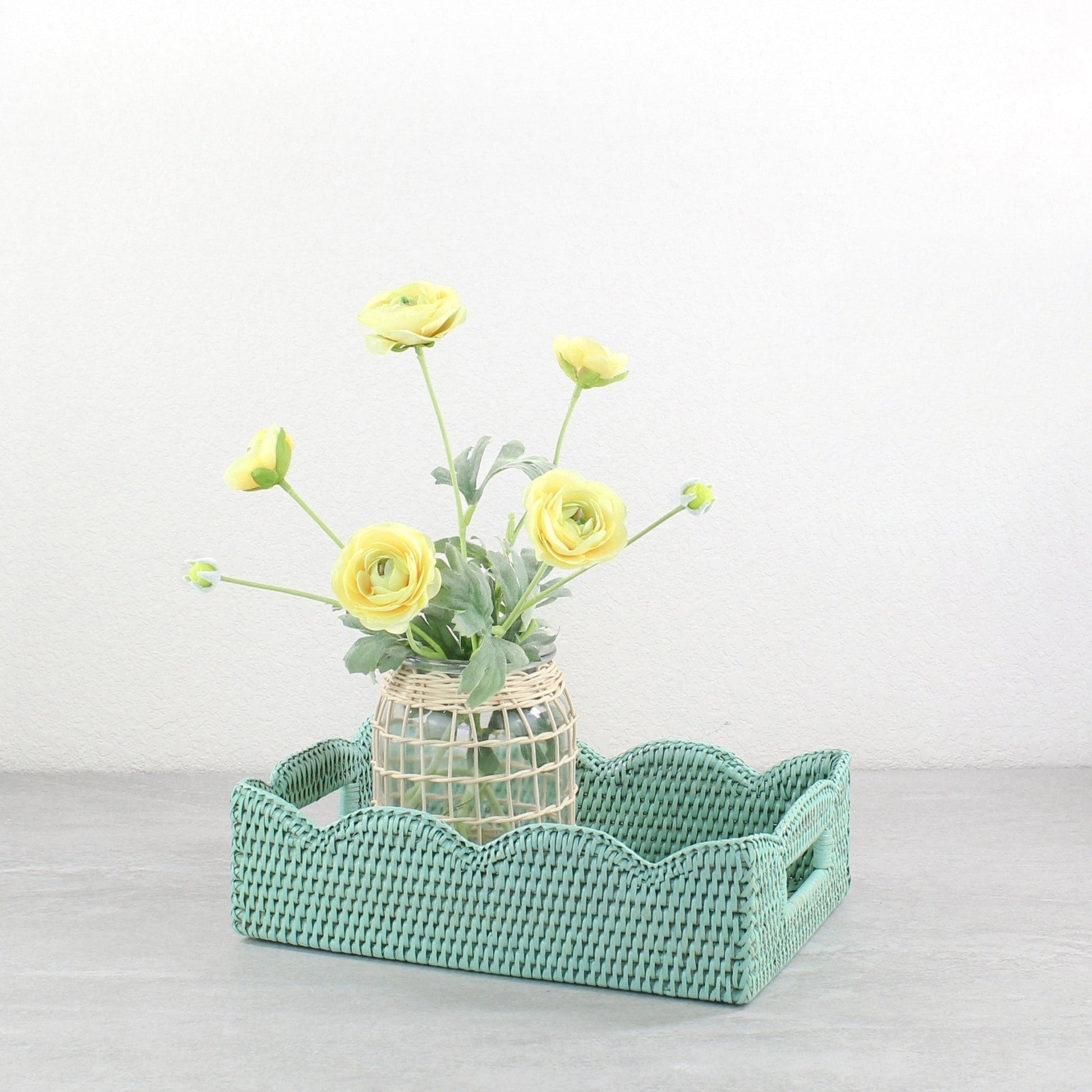 Sage leaf green scalloped rattan tray styled with a woven vase of yellow ranunculus flowers on a neutral tabletop.
