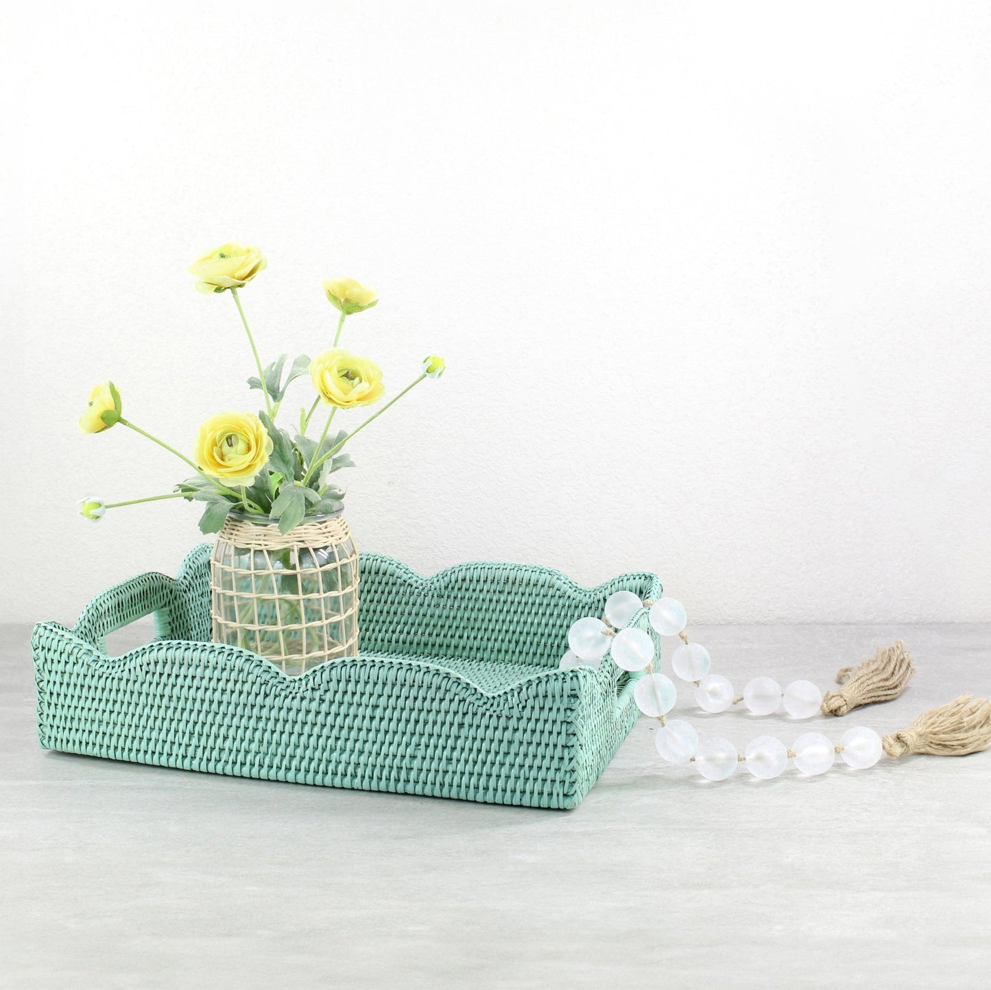 Sage leaf green scalloped rattan tray styled with glass vase, yellow florals, on light grey tabletop.