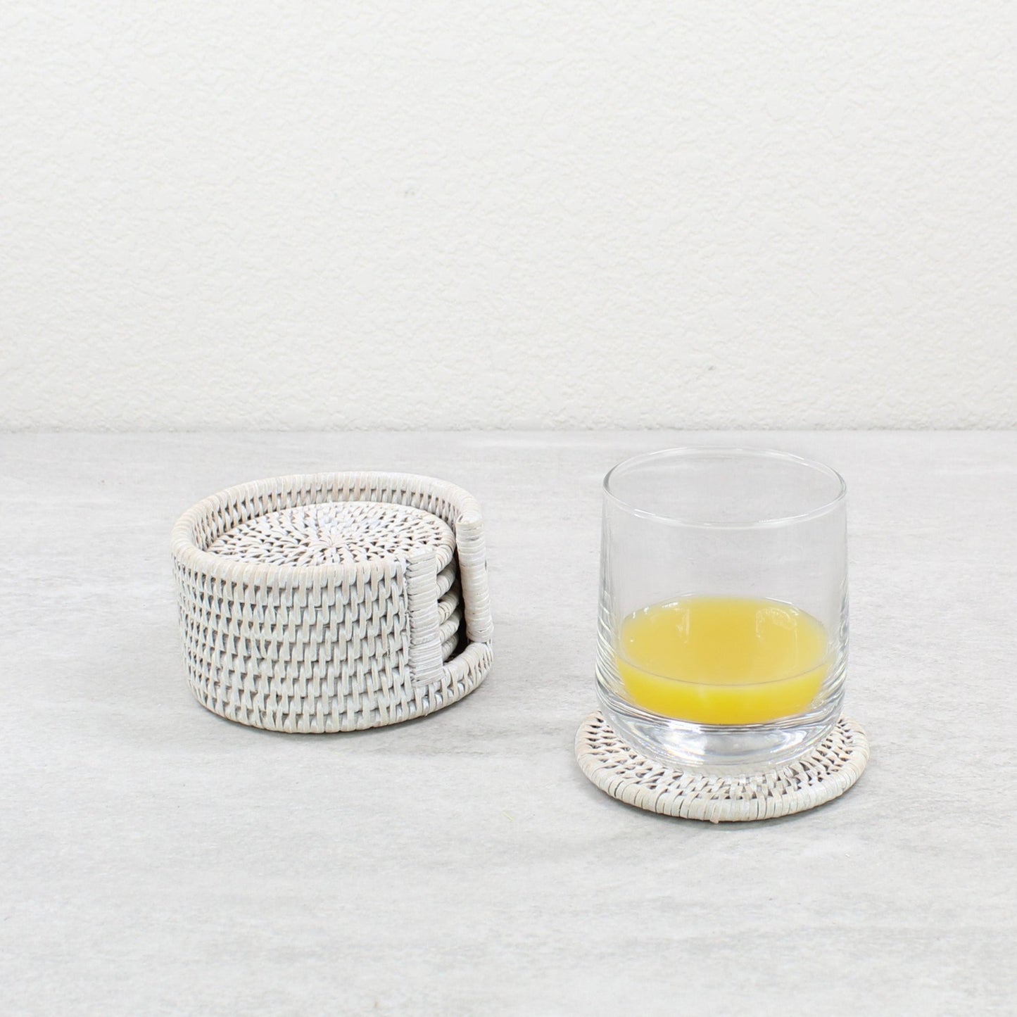 Eco Handcraft handmade whitewash rattan coaster set with holder and a cup of orange juice placed on a grey table.