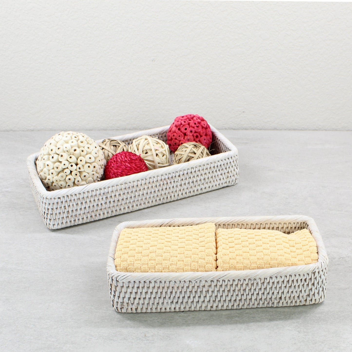 Whitewash rectangular rattan trays holding yellow towels and decorative balls on a gray bathroom counter.