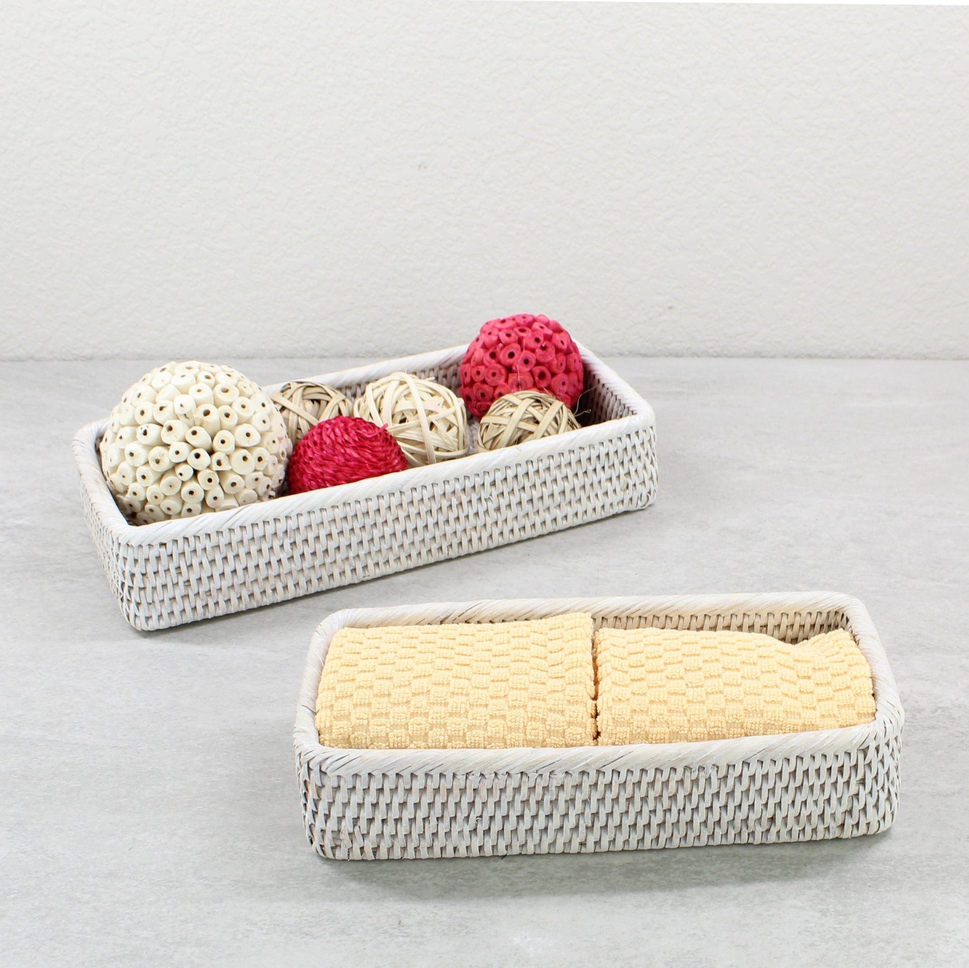Whitewash rectangular rattan trays holding yellow towels and decorative balls on a gray bathroom counter.