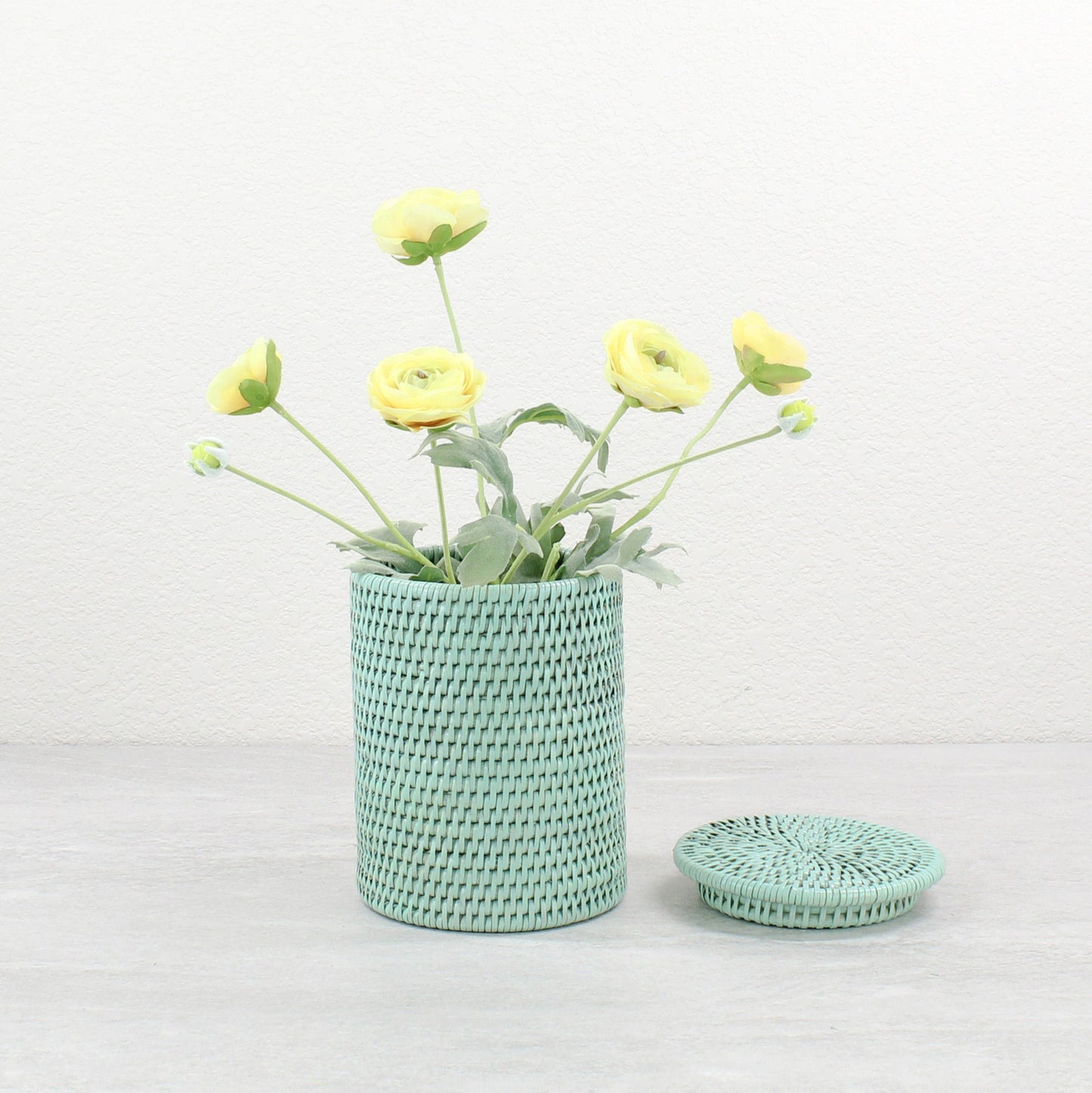 Sage green handwoven rattan canister repurposed as a vase with yellow ranunculus flowers, lid placed beside on light gray surface.