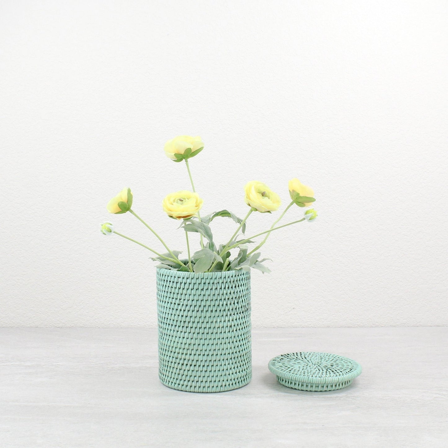 Sage Leaf Green rattan canister with lid, used as a vase for faux yellow blossoms on a light background.