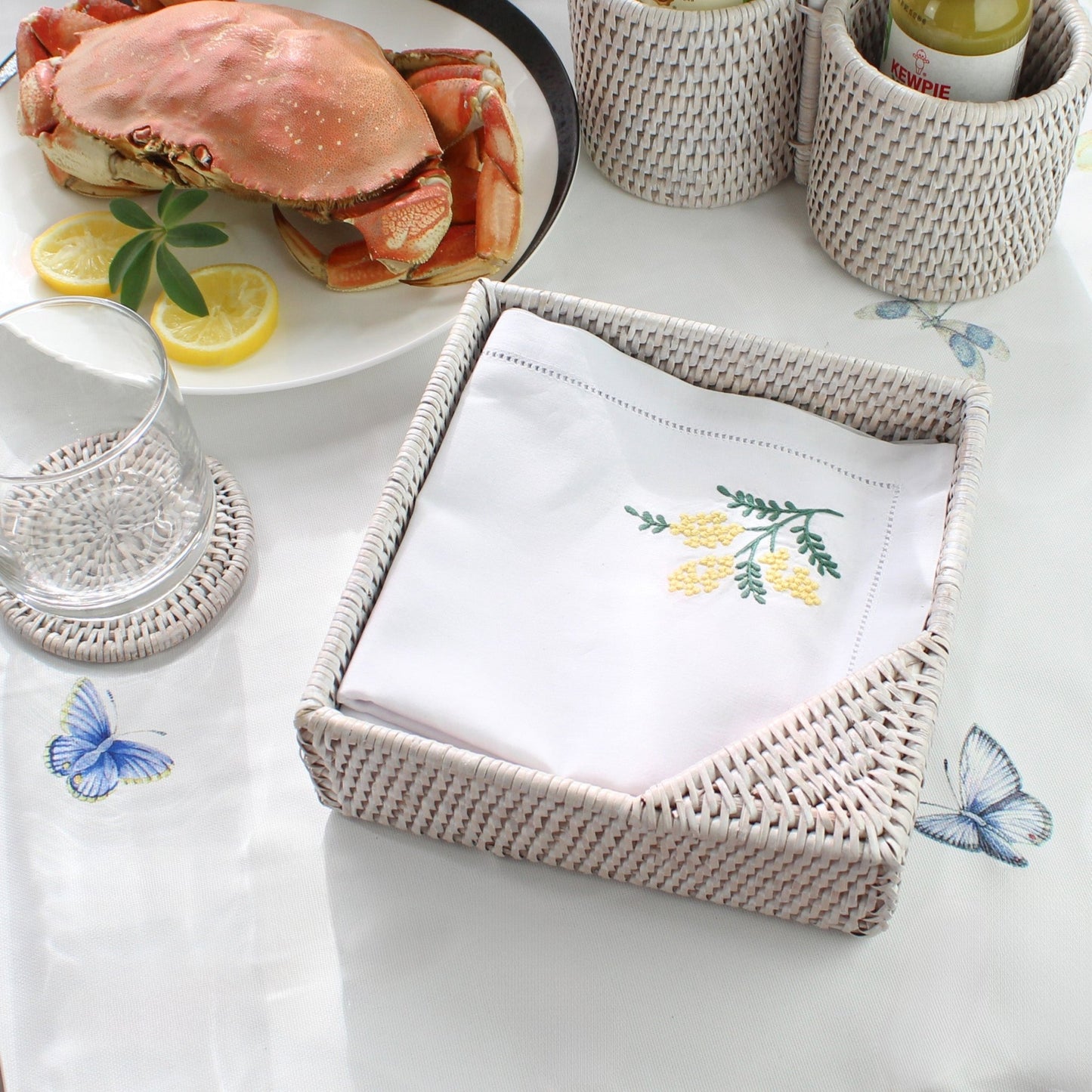 Embroidered napkin in a woven napkin holder with a crab dish and lemon slices in the background.
