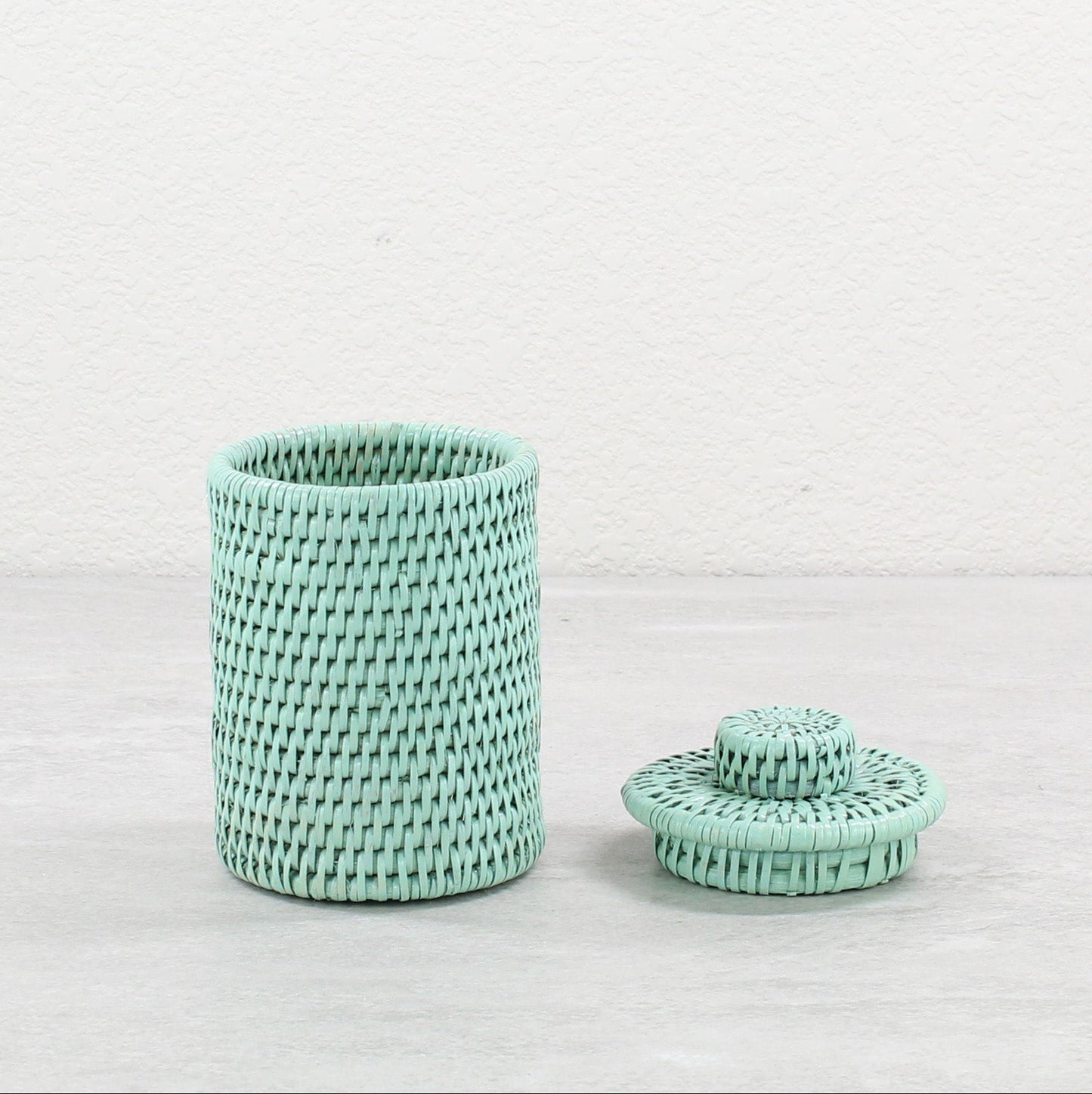 Handwoven sage green rattan canister with lid, lid placed beside the canister, displayed on a neutral surface.