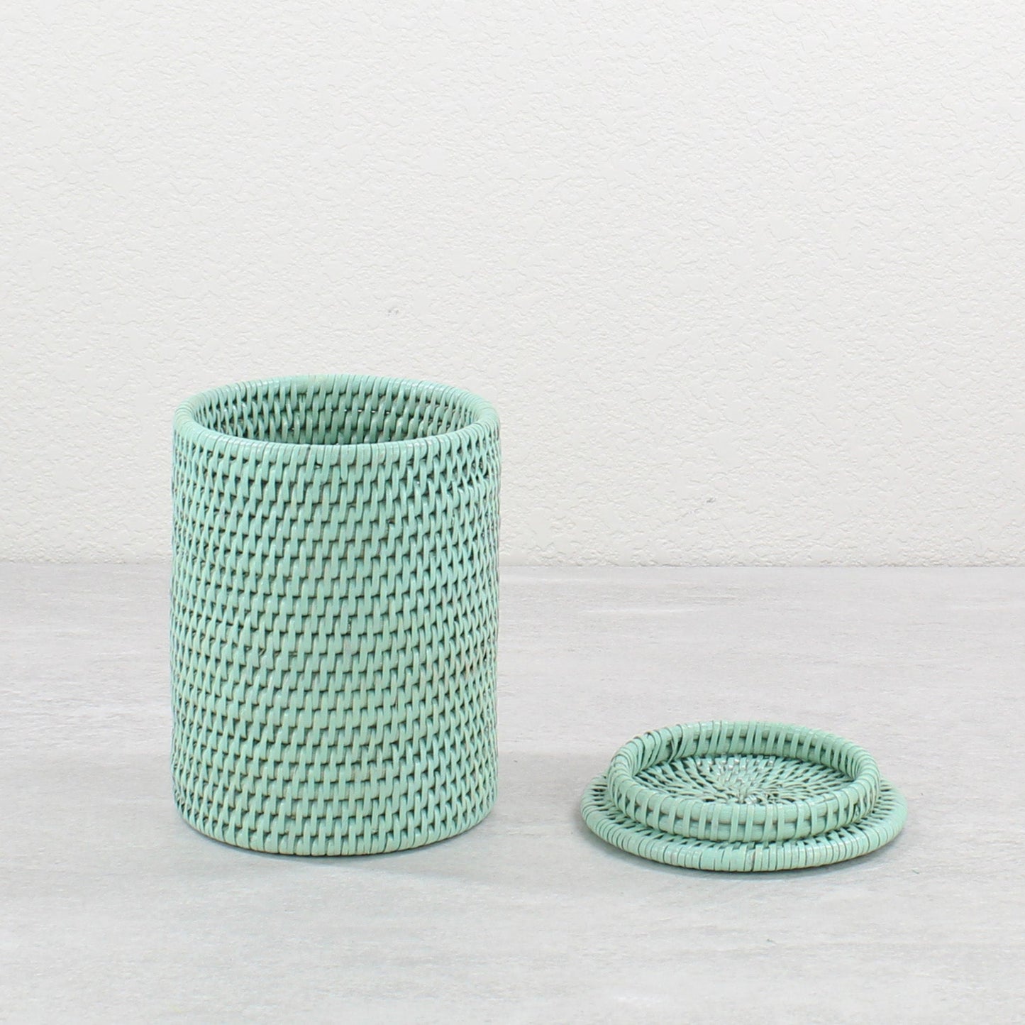 Sage green handwoven rattan canister, lid placed beside on light gray surface.