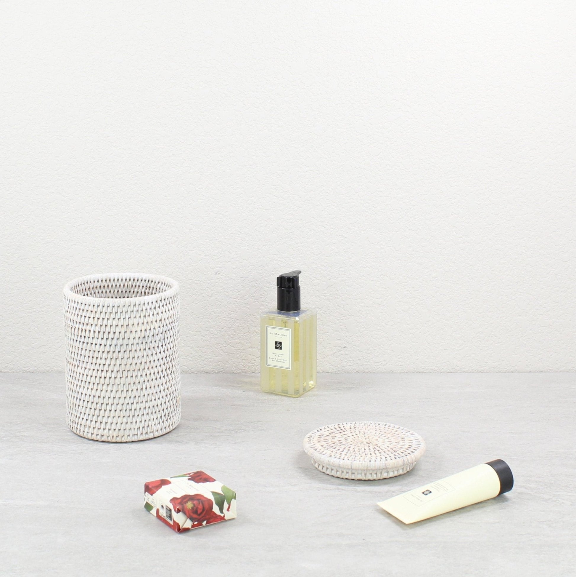Whitewash rattan canister with lid styled with perfume, floral soap, and lotion on a light gray bathroom surface.