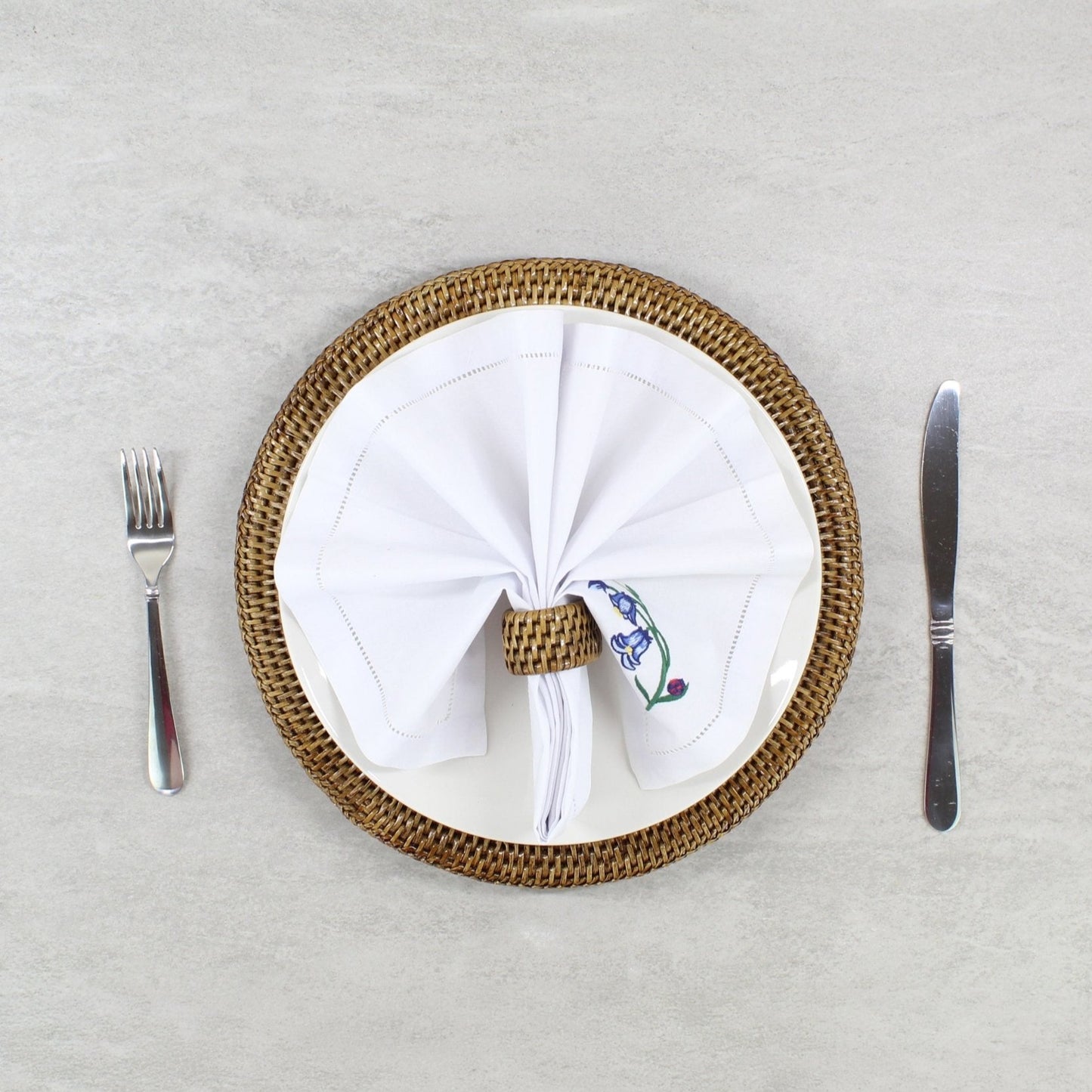 Eco Handcraft rattan placemat and napkin ring styled with white embroidered napkin and modern flatware on a minimalist table.