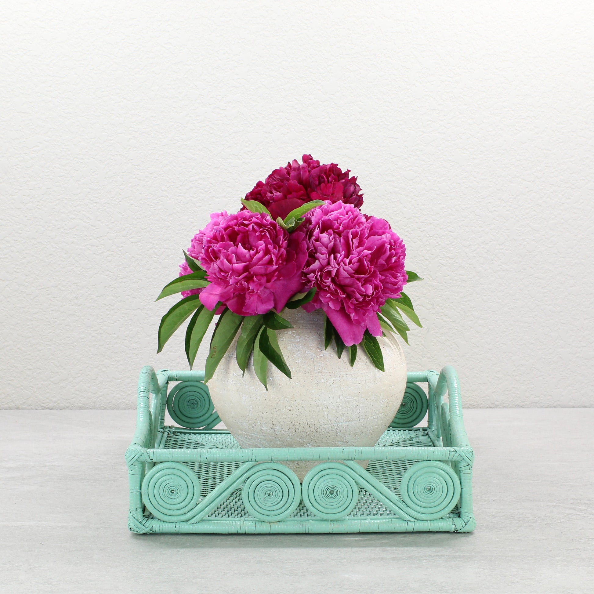Sage Leaf Green rattan tray with swirl design holding a cream vase of vibrant pink peonies for modern interior styling.