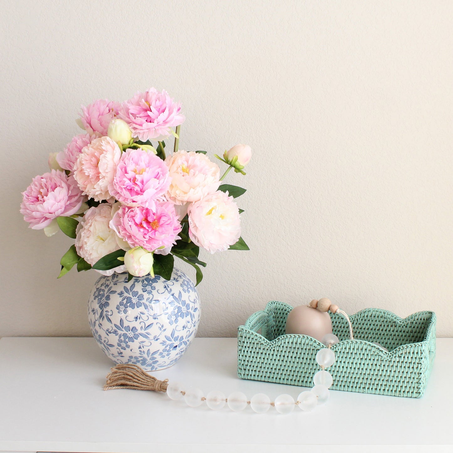 Sage leaf green small scalloped rattan tray with pink peony bouquet in blue vase, styled on a white dresser for elegant home decor.