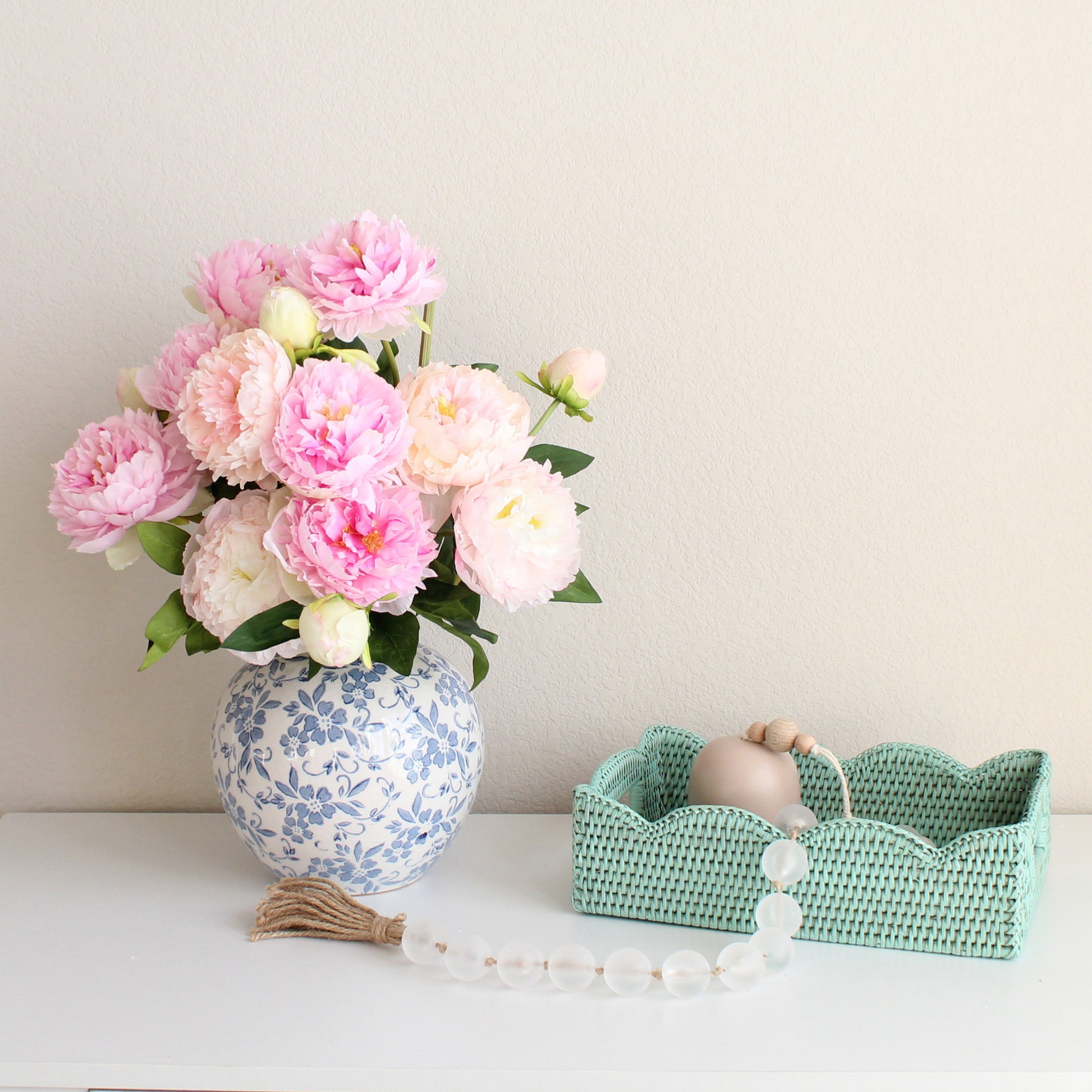 Sage leaf green small scalloped rattan tray with pink peony bouquet in blue vase, styled on a white dresser for elegant home decor.
