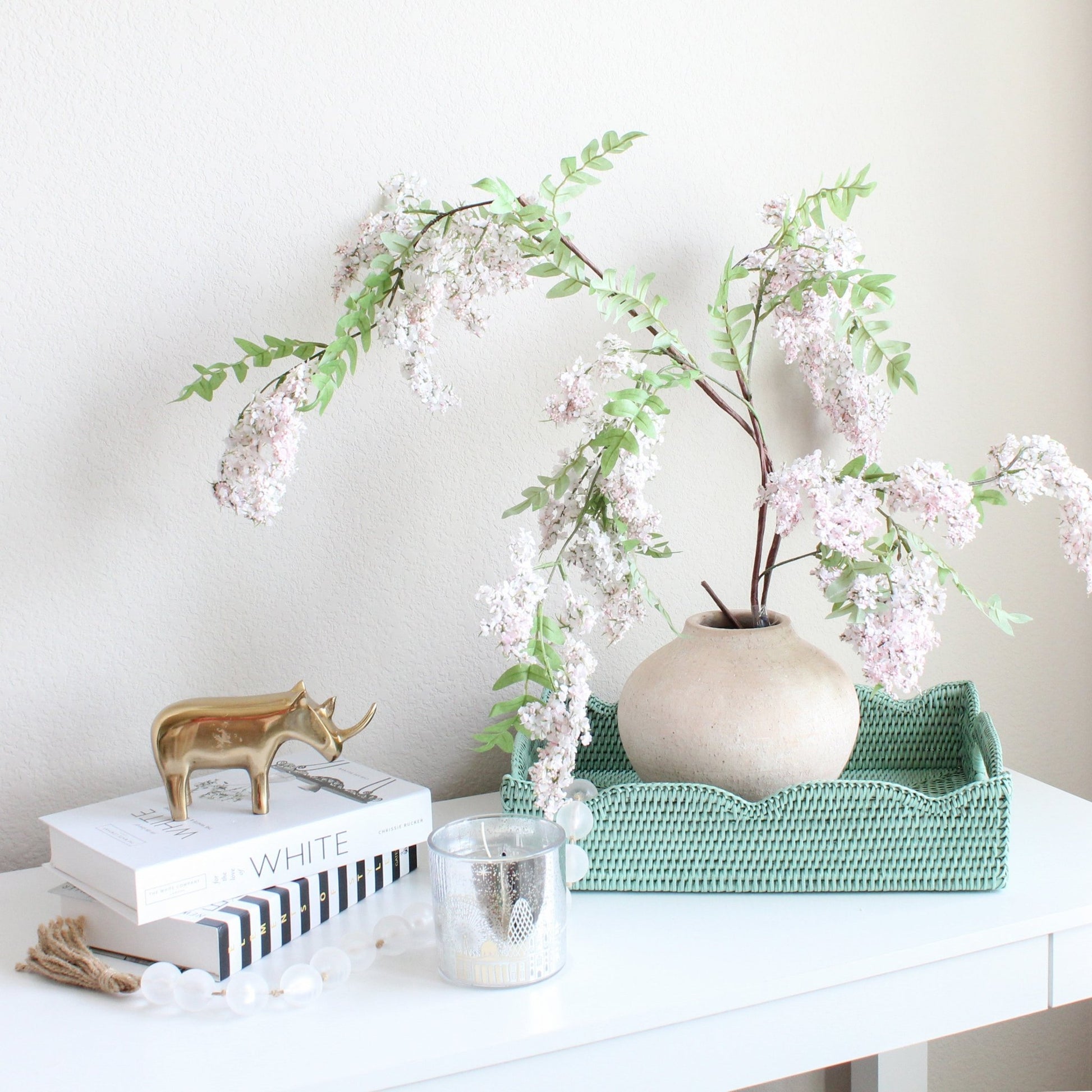 Sage leaf green scalloped rattan tray styled with neutral vase, pink florals, books, and gold accent on white tabletop.