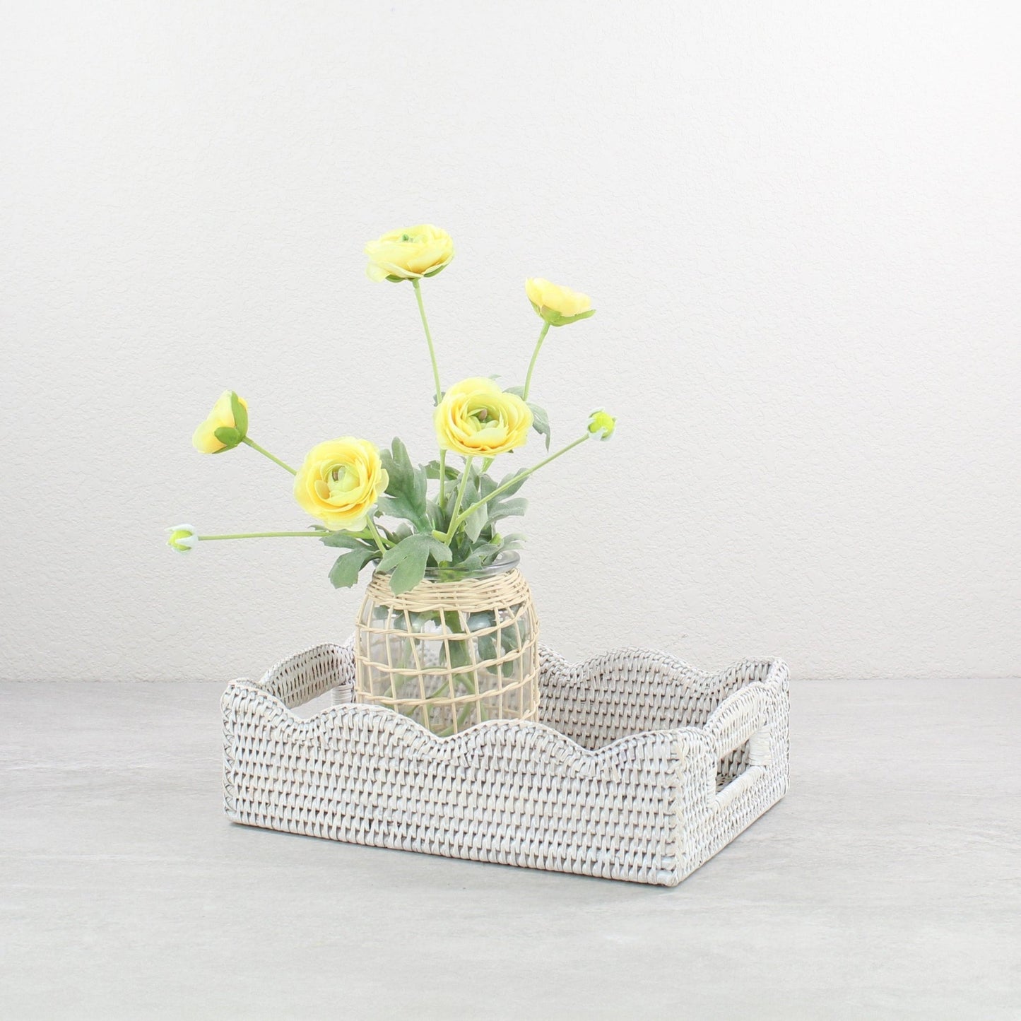 Decorative scalloped whitewash rattan tray with a vase of yellow flowers on a light gray background