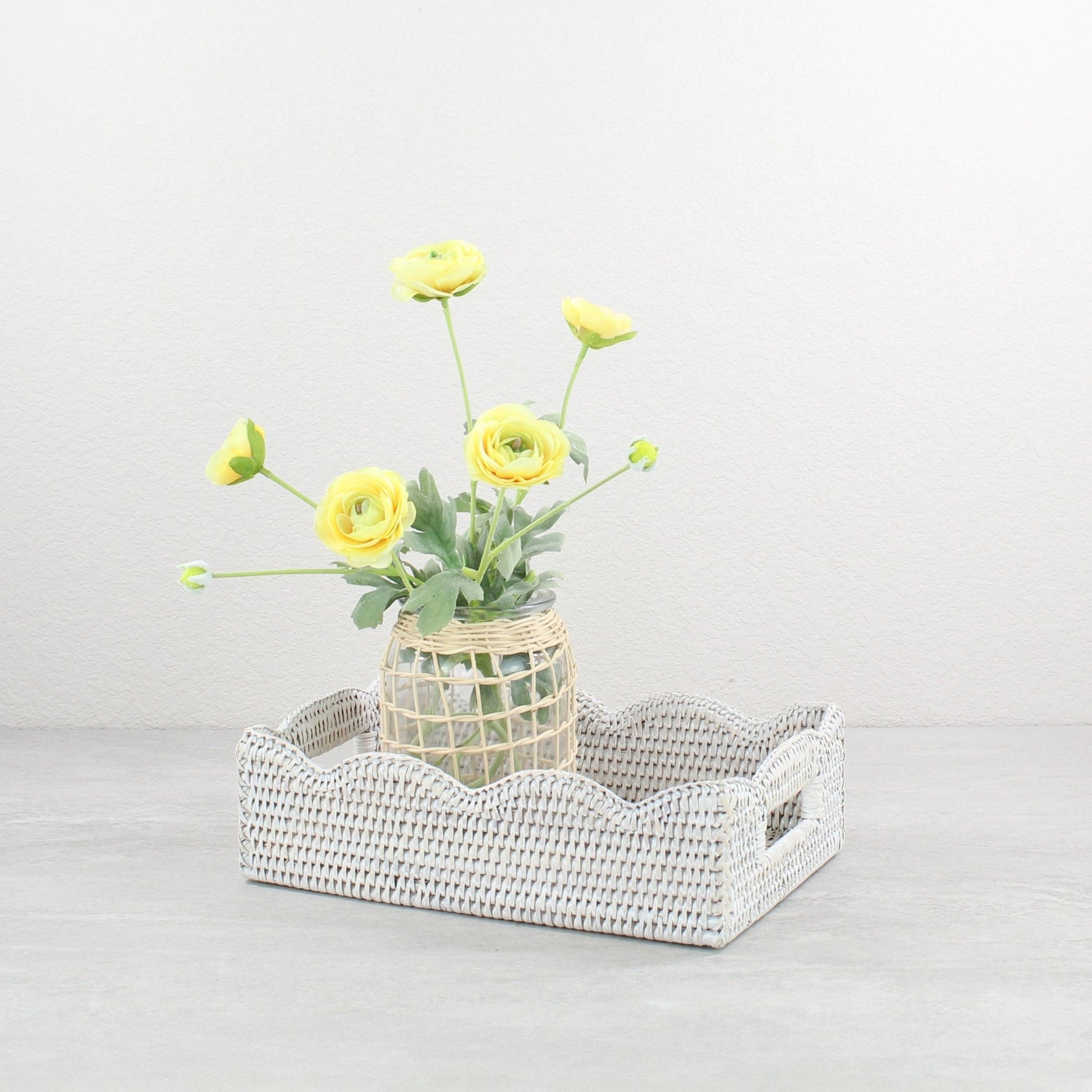 Decorative scalloped whitewash rattan tray with a vase of yellow flowers on a light gray background