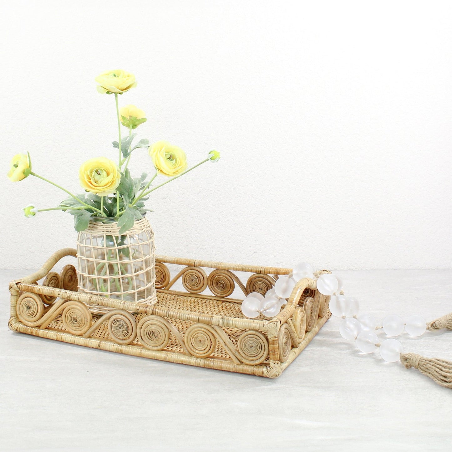 Natural rectangle rattan tray styled with yellow ranunculus flowers in a woven vase and frosted bead garland.