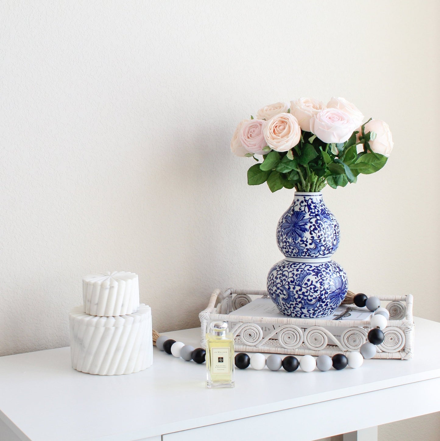 White swirly rattan tray styled with blue floral vase, roses, and perfume on white table for chic modern home décor.