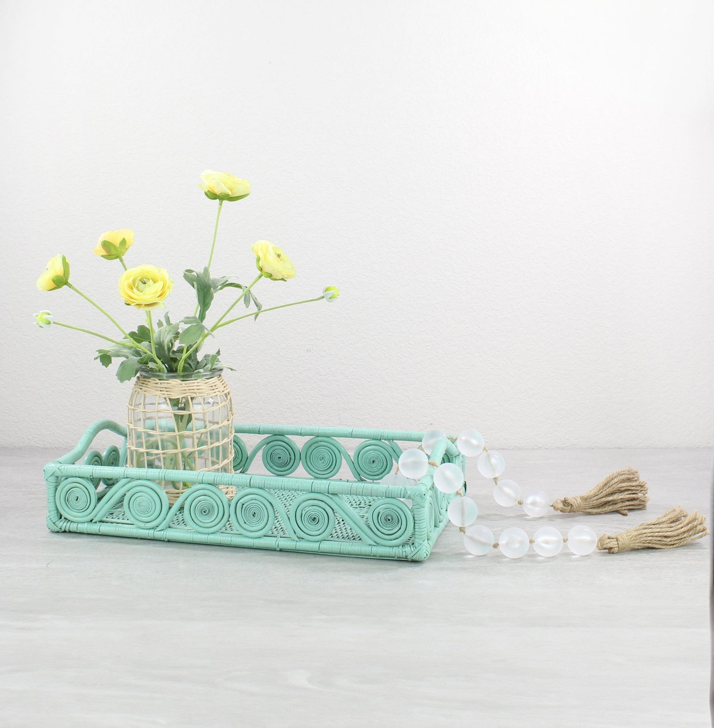 Turquoise swirly rattan tray styled with yellow flowers in a woven vase and glass bead garland on a gray tabletop.