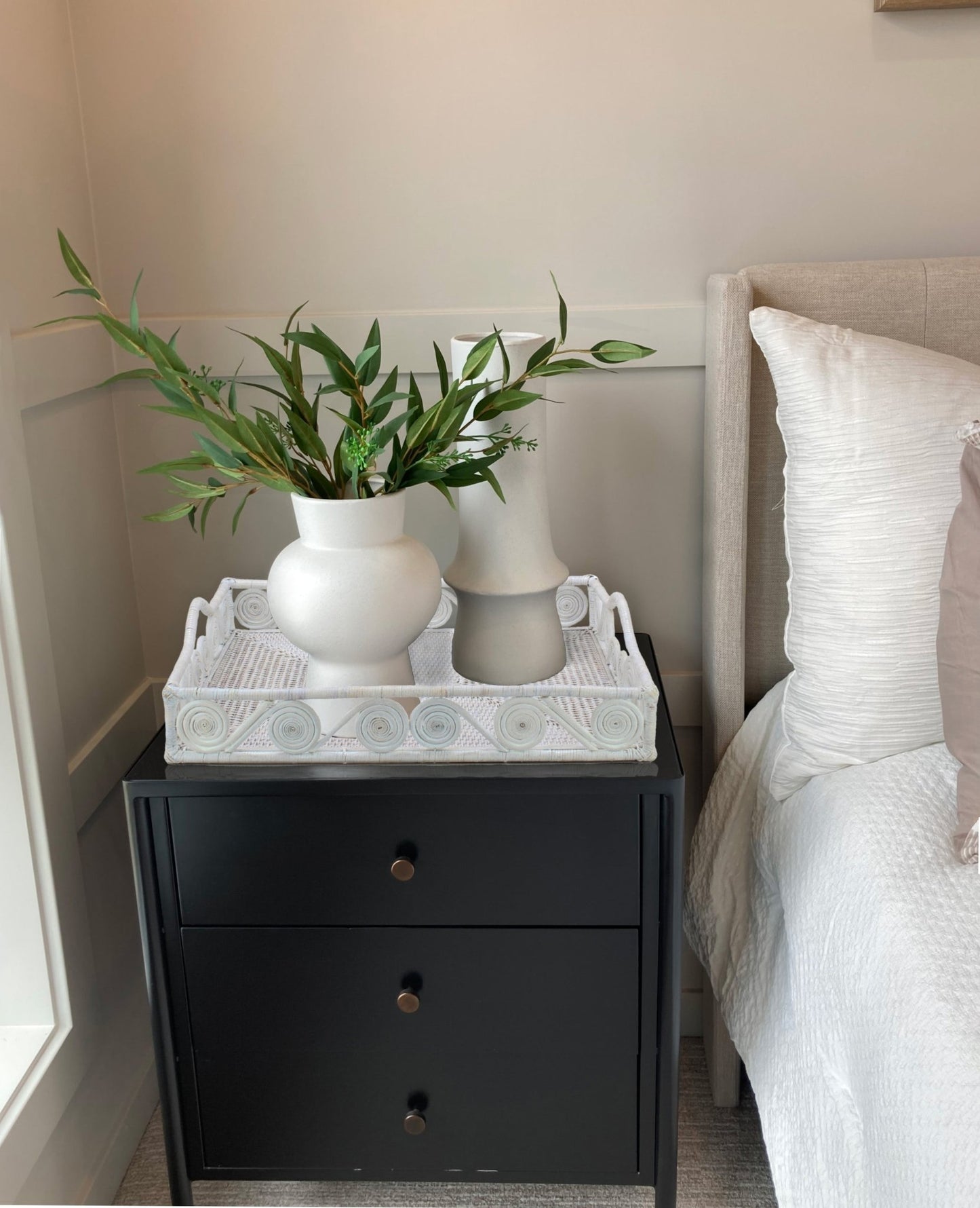 Swirly white rattan tray with vases on black nightstand beside bed, adding texture to a cozy modern bedroom corner.