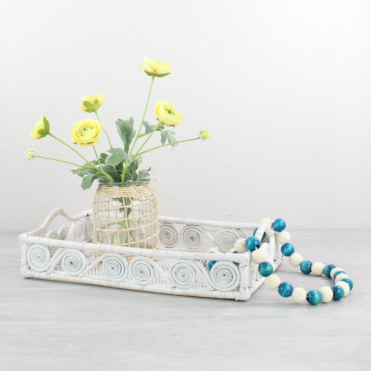 Whitewash swirly rattan tray styled with yellow flowers in a woven vase and blue bead garland on a gray tabletop.