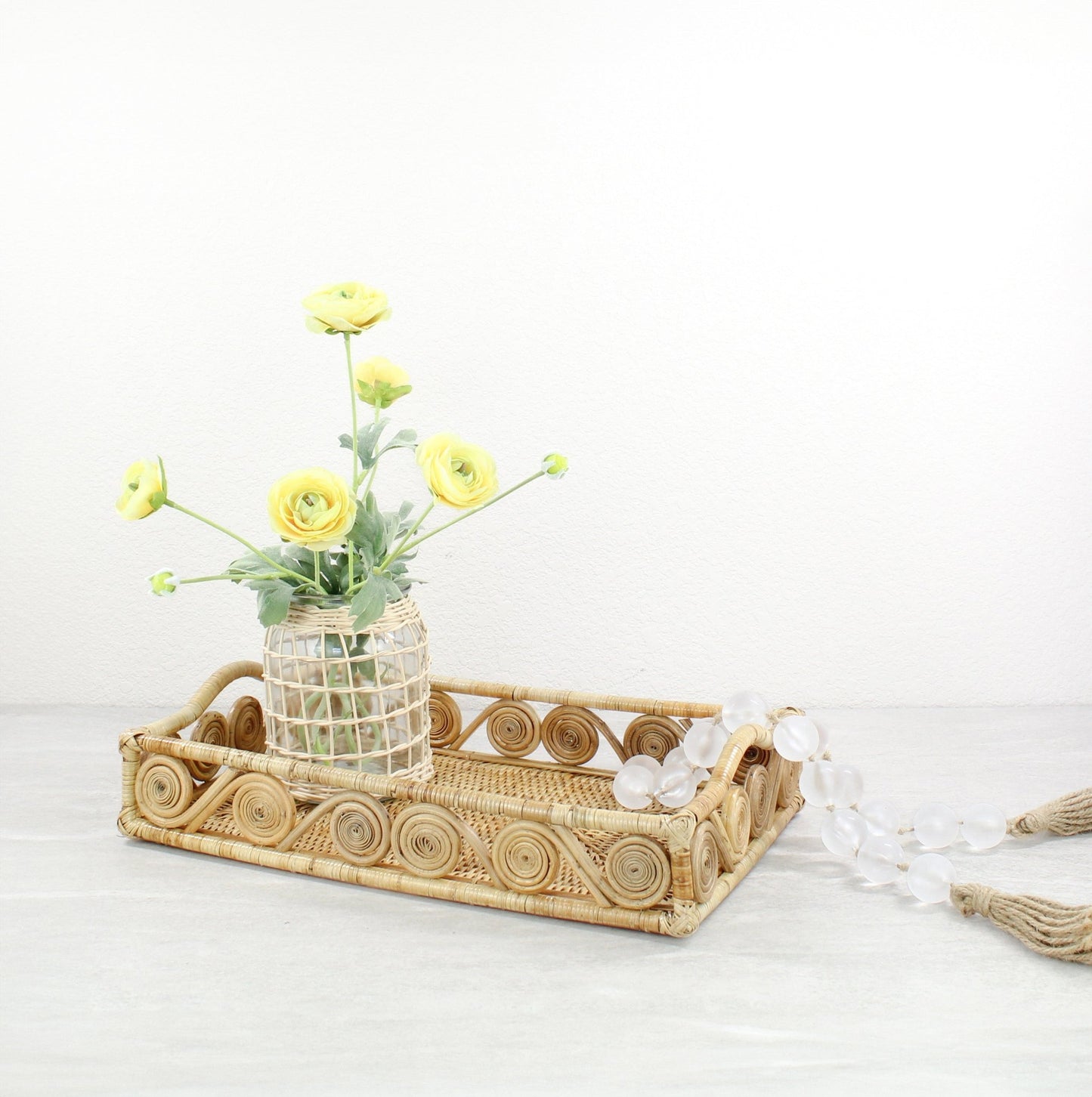 Natural swirly rattan tray styled with yellow flowers in a woven vase and glass bead garland on a gray tabletop.