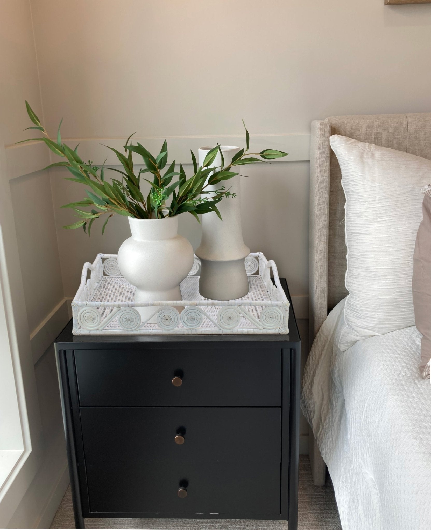 Whitewash rattan tray with swirl design styled on a black nightstand beside a bed, holding modern white ceramic vases.