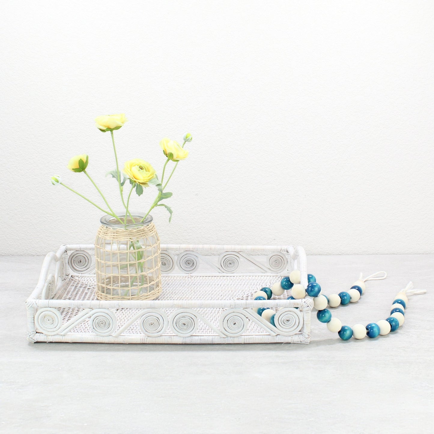 Whitewash rattan tray styled with yellow ranunculus flowers and blue decorative beads on a neutral tabletop.
