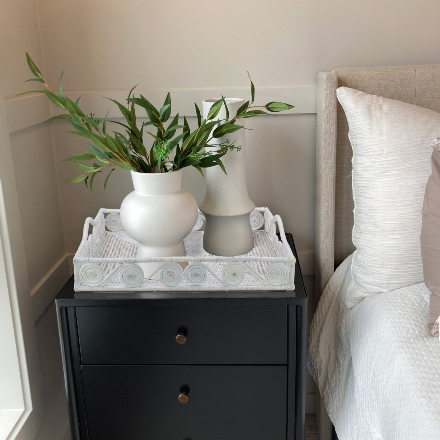 White rattan tray with swirl design styled on a black nightstand beside a bed, holding modern white ceramic vases.